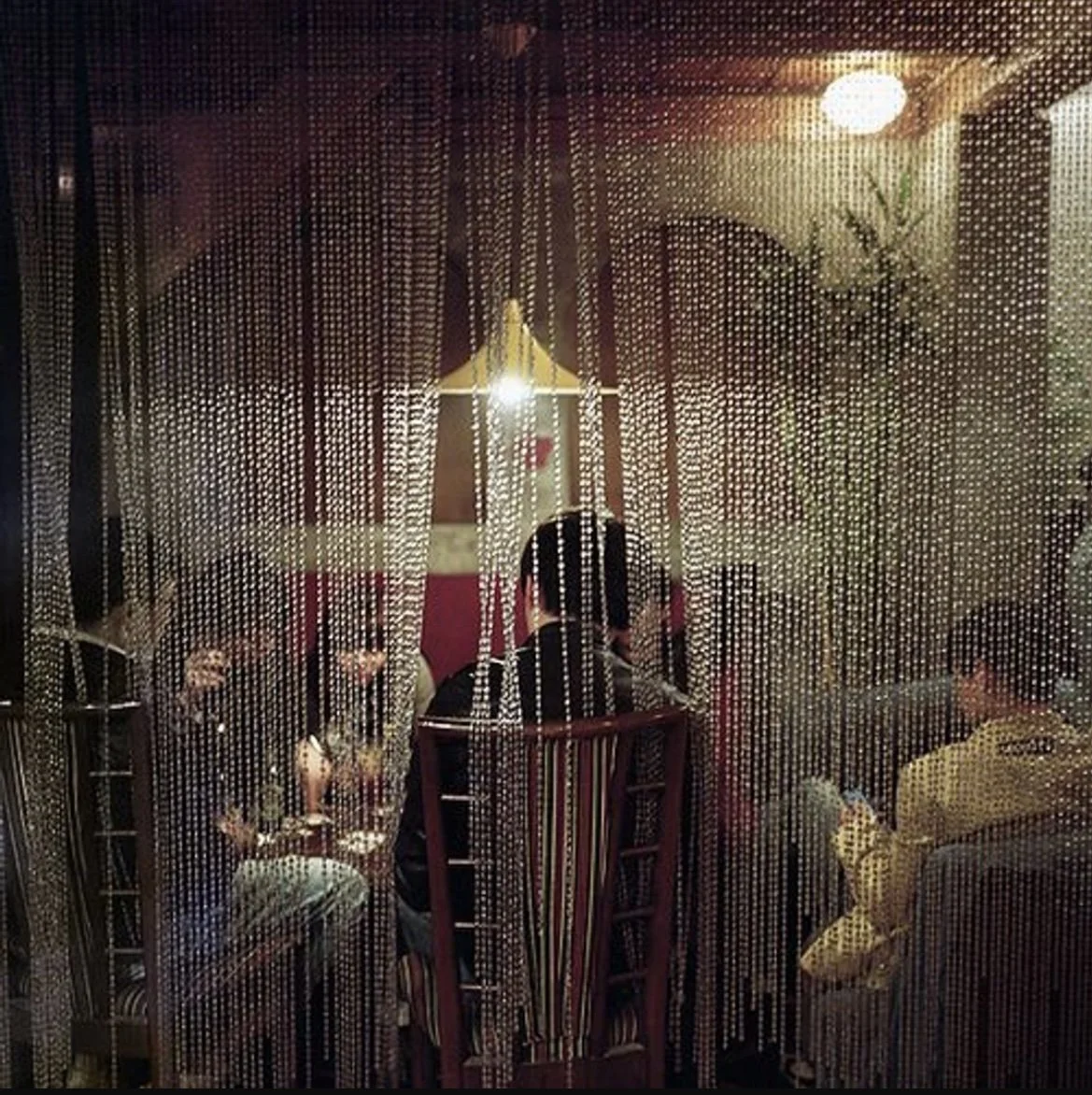 View through a beaded curtain into a dimly lit restaurant where four people are seated at a table. The table has two glasses of wine and several plates. The scene is illuminated by a wall-mounted lamp and a ceiling light, with a decorative plant visible on the right.