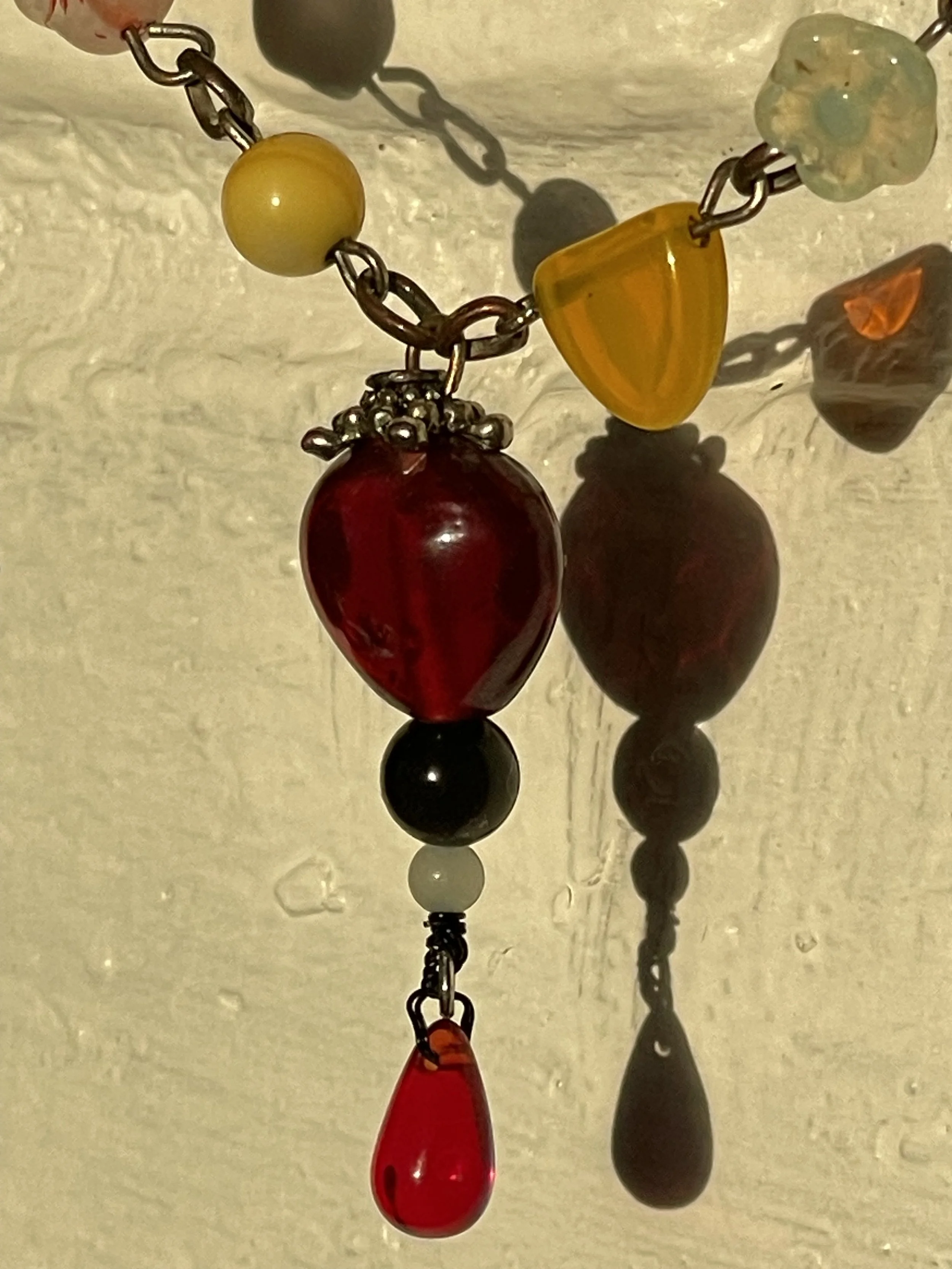 Close-up of a colorful beaded necklace with red, yellow, black, and multicolored beads hanging against a beige background, casting reflections and shadows.