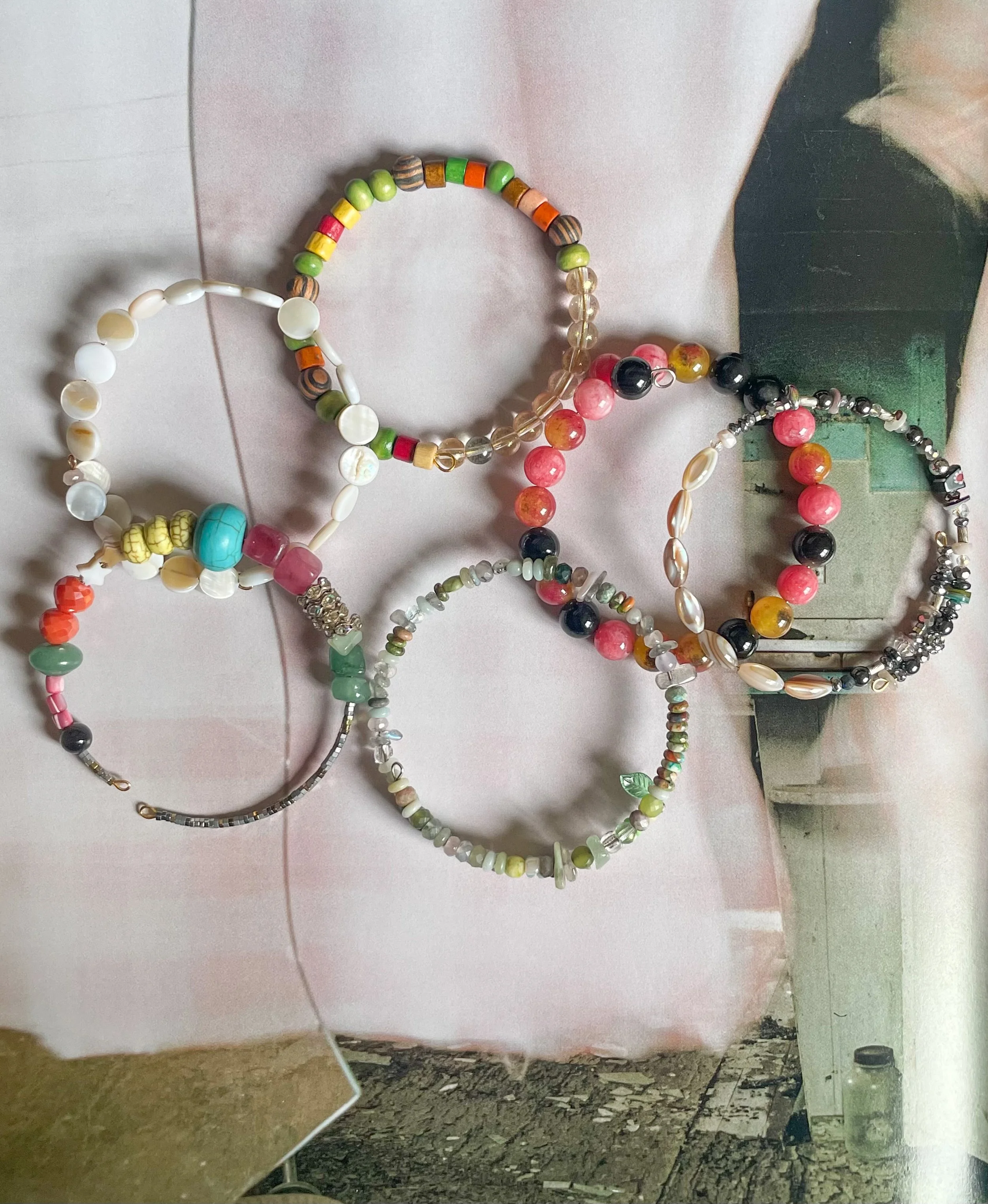 Collection of colorful beaded necklaces laid out on a pink surface, with a mirror reflecting part of the display and a plastic bottle on the ground.