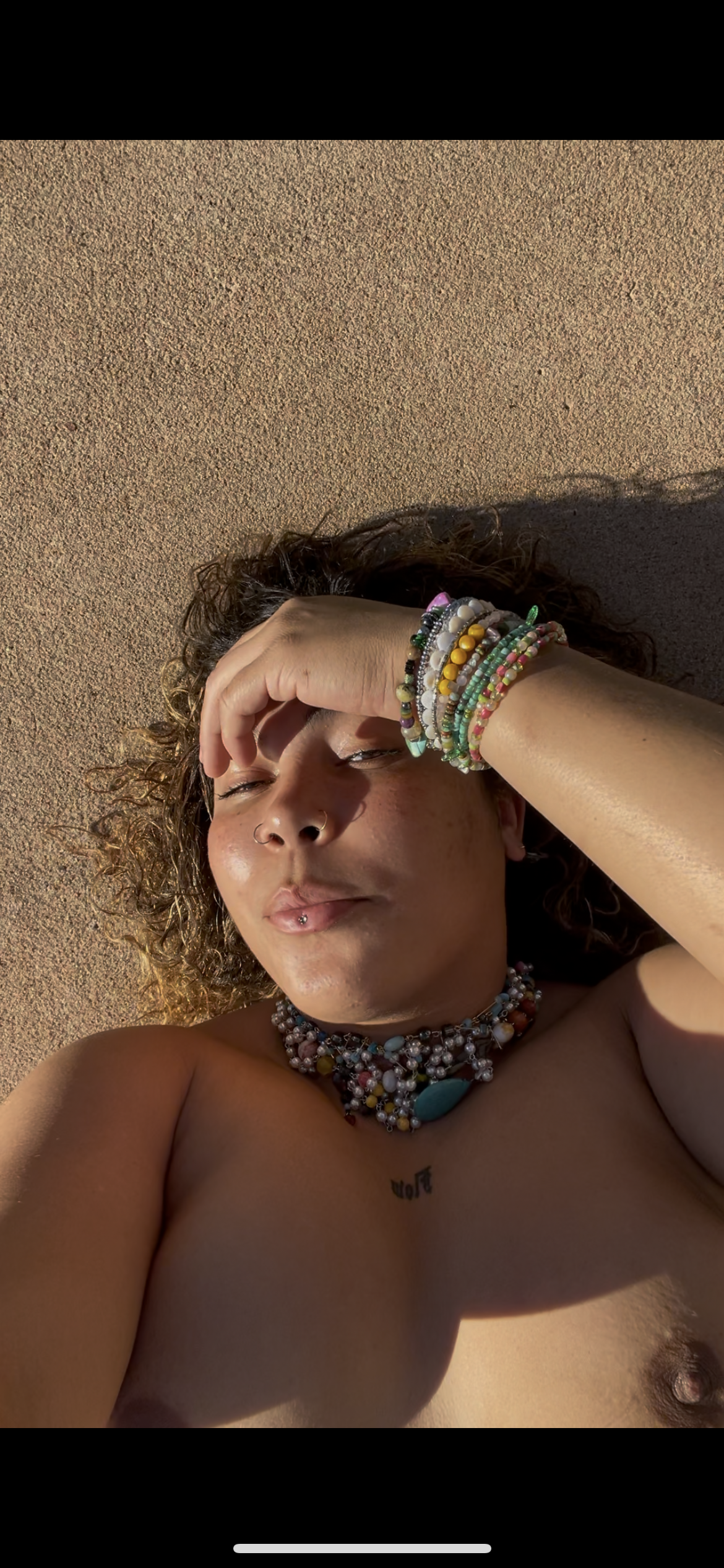 A woman with curly hair lying on sandy ground, wearing multiple colorful beaded bracelets, a beaded necklace, a nose ring, and a lip piercing, with her hand resting on her forehead.