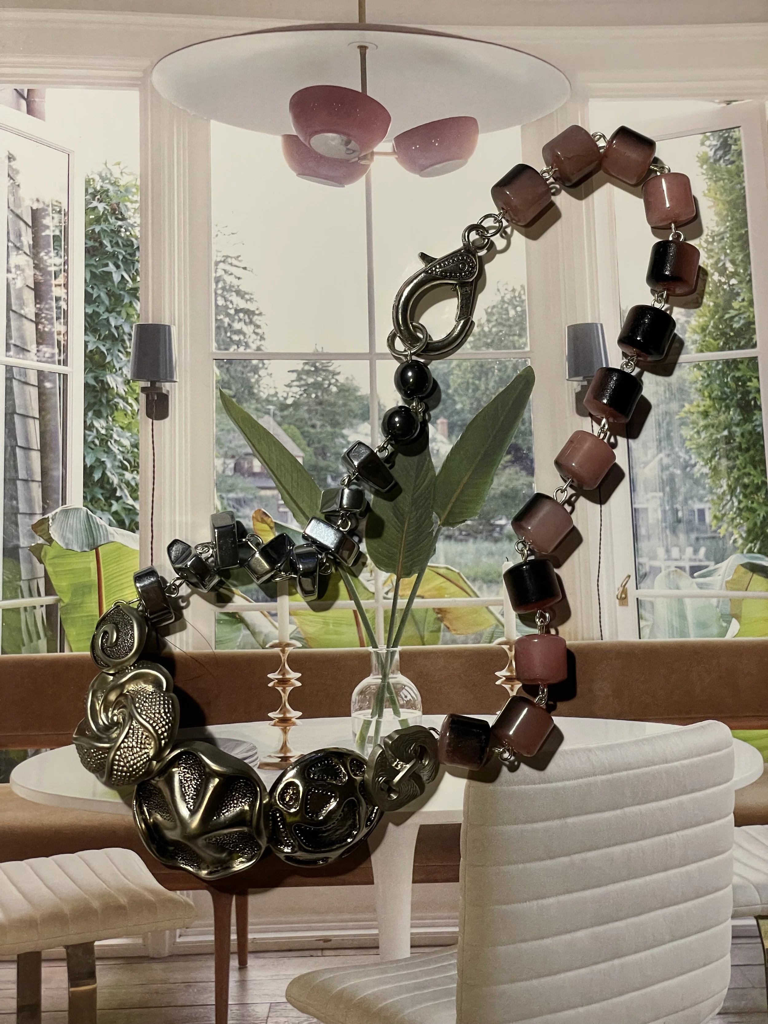 A round white table with white tablecloth, adorned with black, silver, and pink beaded jewelry, including a large black and silver bracelet, a pearl necklace, and multi-colored beaded necklace. A clear glass vase with green leaves is in the background near a window with a garden view.
