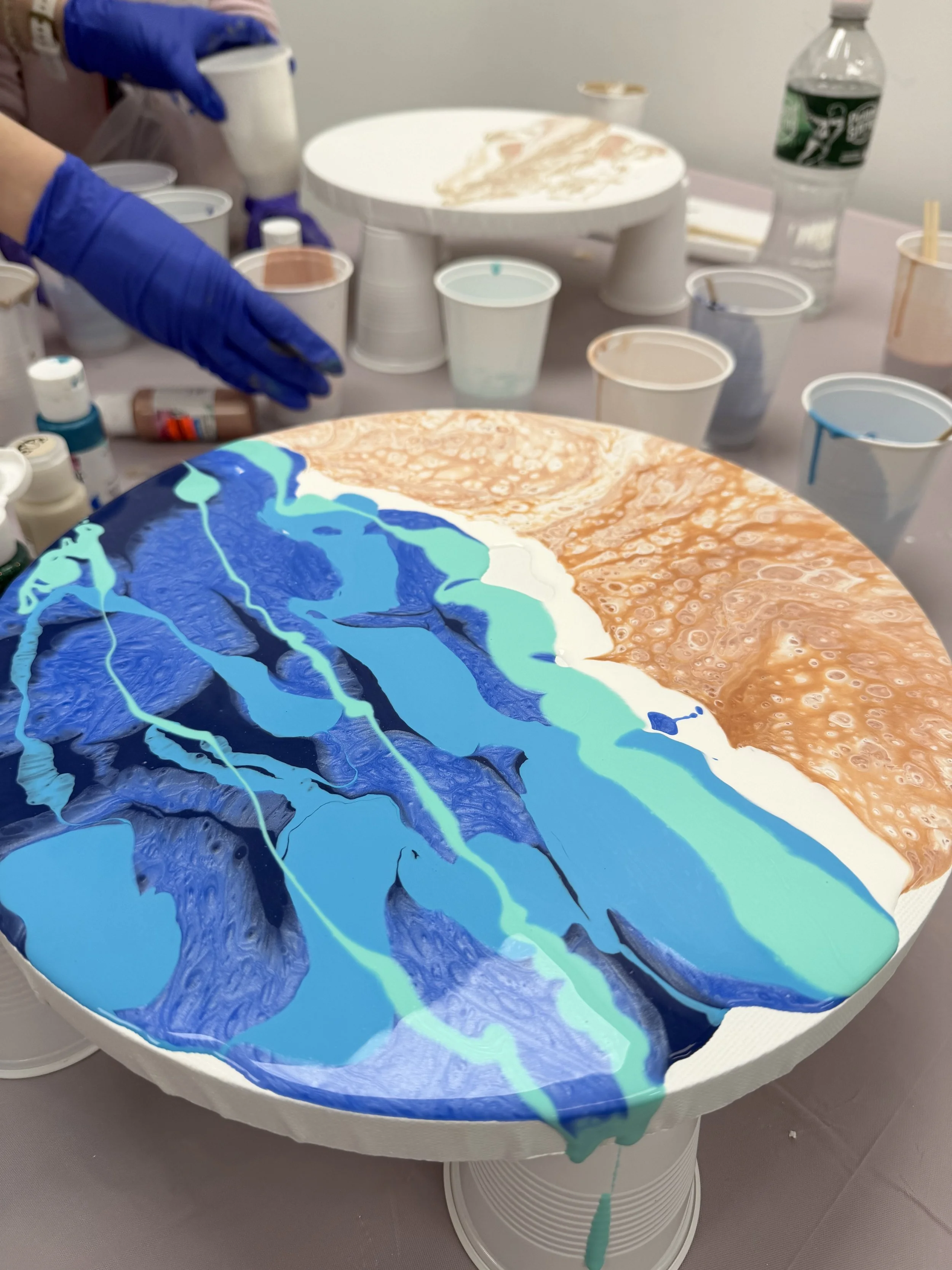 Circular canvas with abstract drip painting in blue, teal, white, and copper colors, with various cups and art supplies on surrounding table.