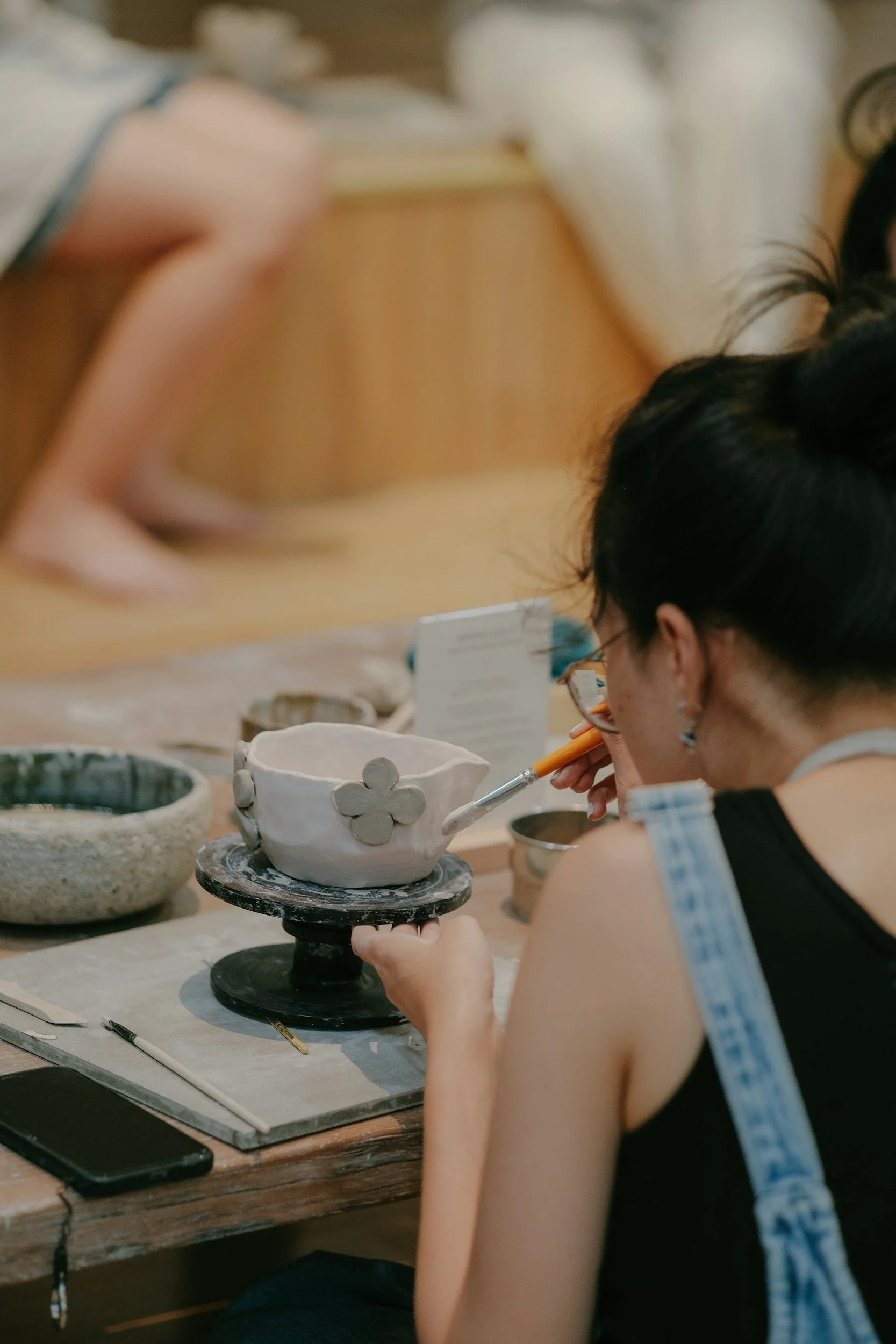 A woman is painting a small, white ceramic bowl decorated with flower shapes at a pottery studio. In the background, a person is squatting on the floor.