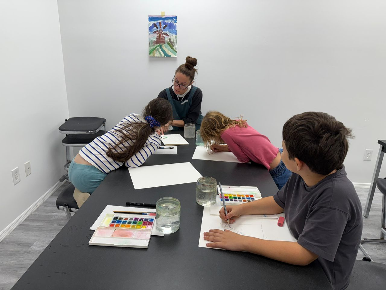 A woman teaching art to three children at a table in a classroom with white walls and a colorful painting hanging on the wall.