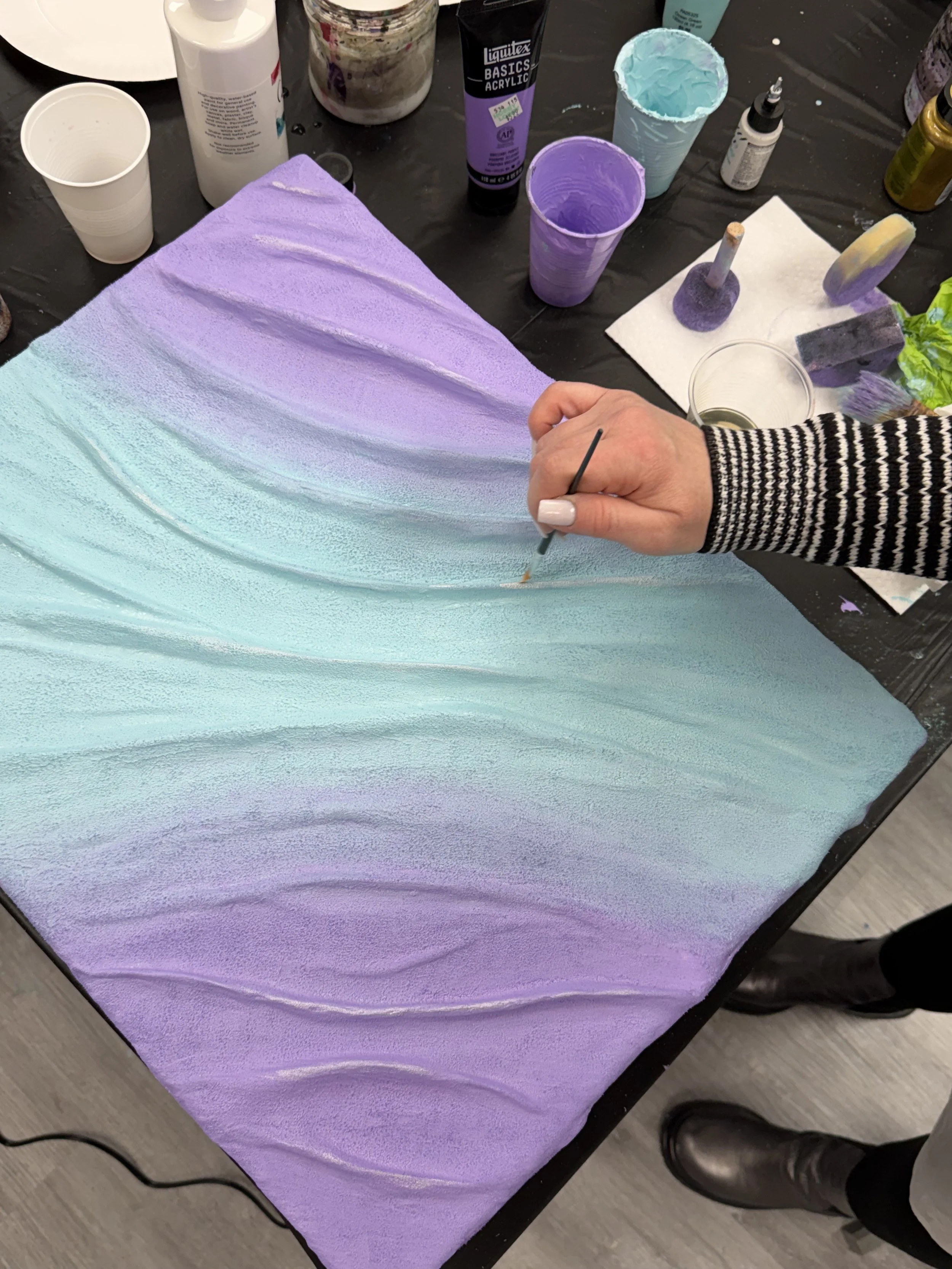 Person painting a textured, abstract pastel-colored canvas with shades of purple, blue, and green at an art studio.