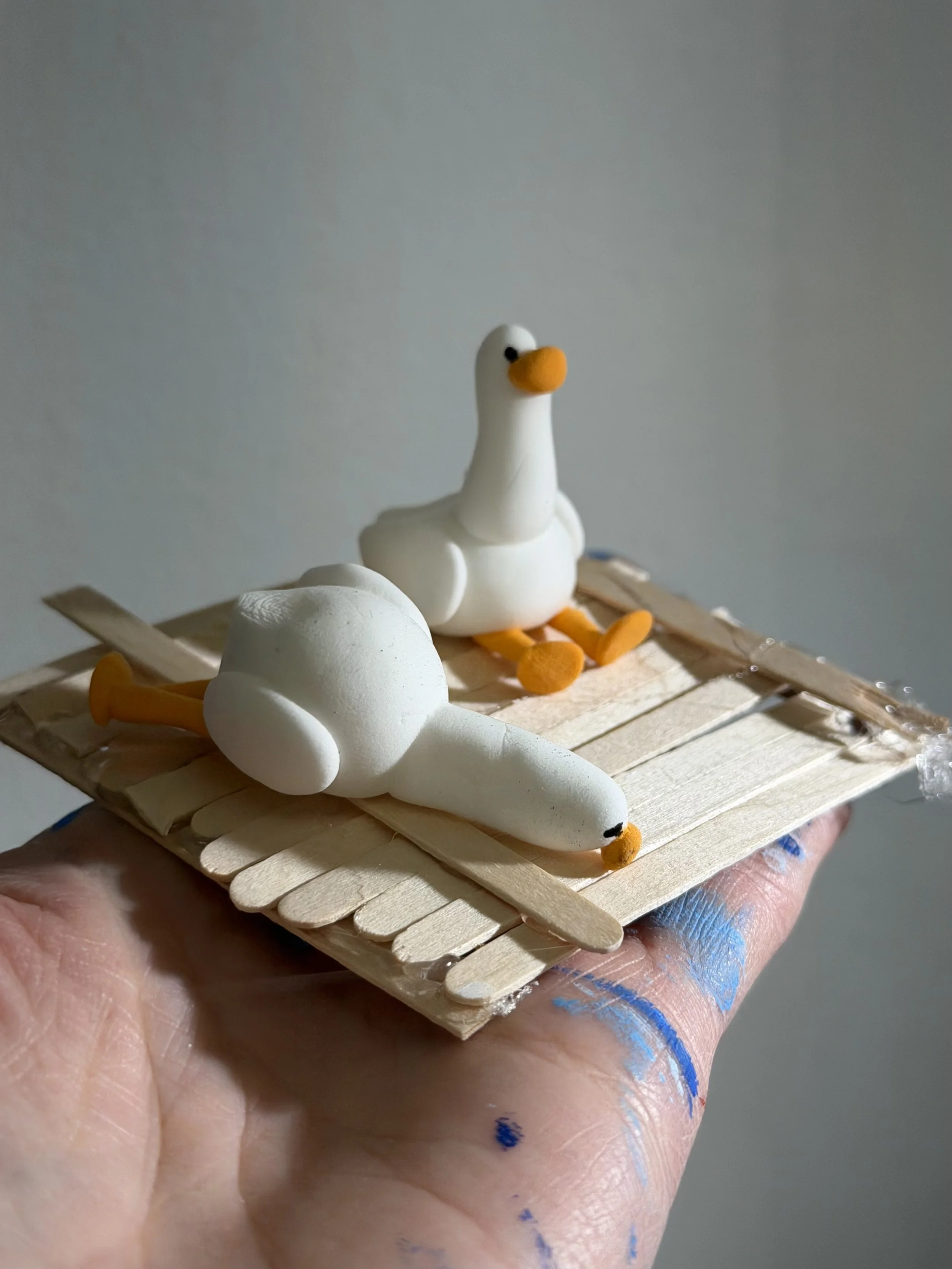 Hand holding a small wooden platform with two foam ducks, one sitting upright and the other lying on its side, resembling a duck family.