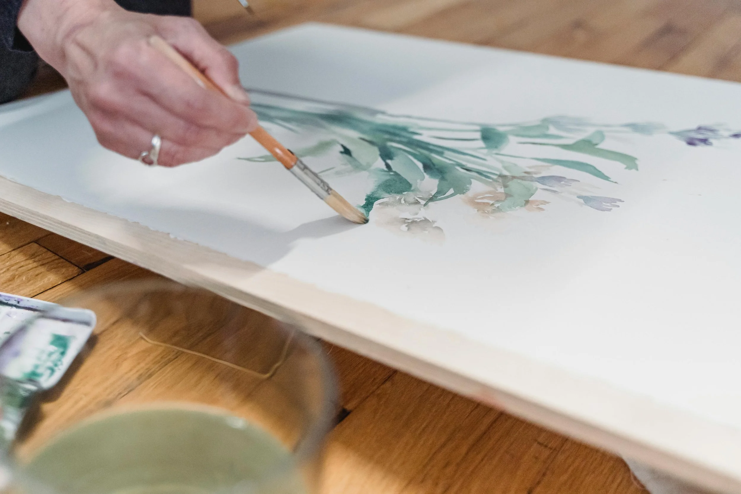 A person painting a floral watercolor on paper, with a brush in hand, placed on a wooden table.