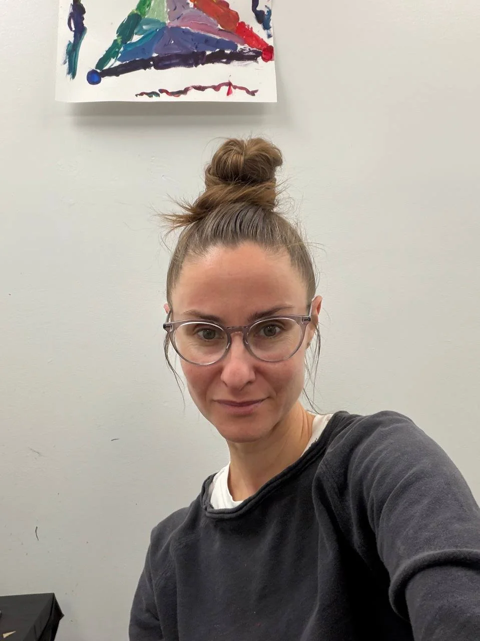 A woman with glasses and light skin taking a selfie in front of a wall with a colorful abstract painting.