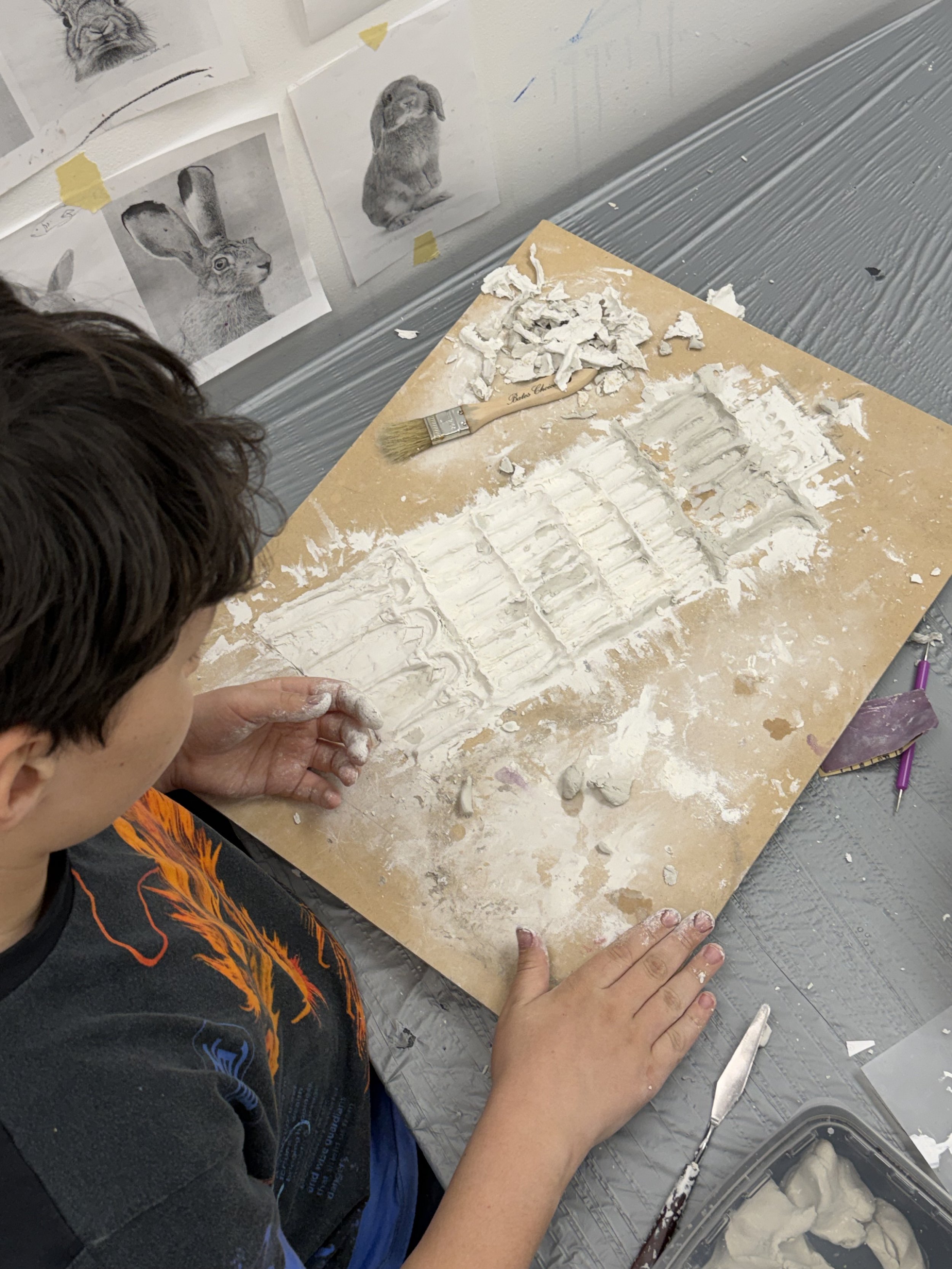 Child working on a clay sculpture or carving on a wooden board, with pictures of animals taped to the wall behind them.