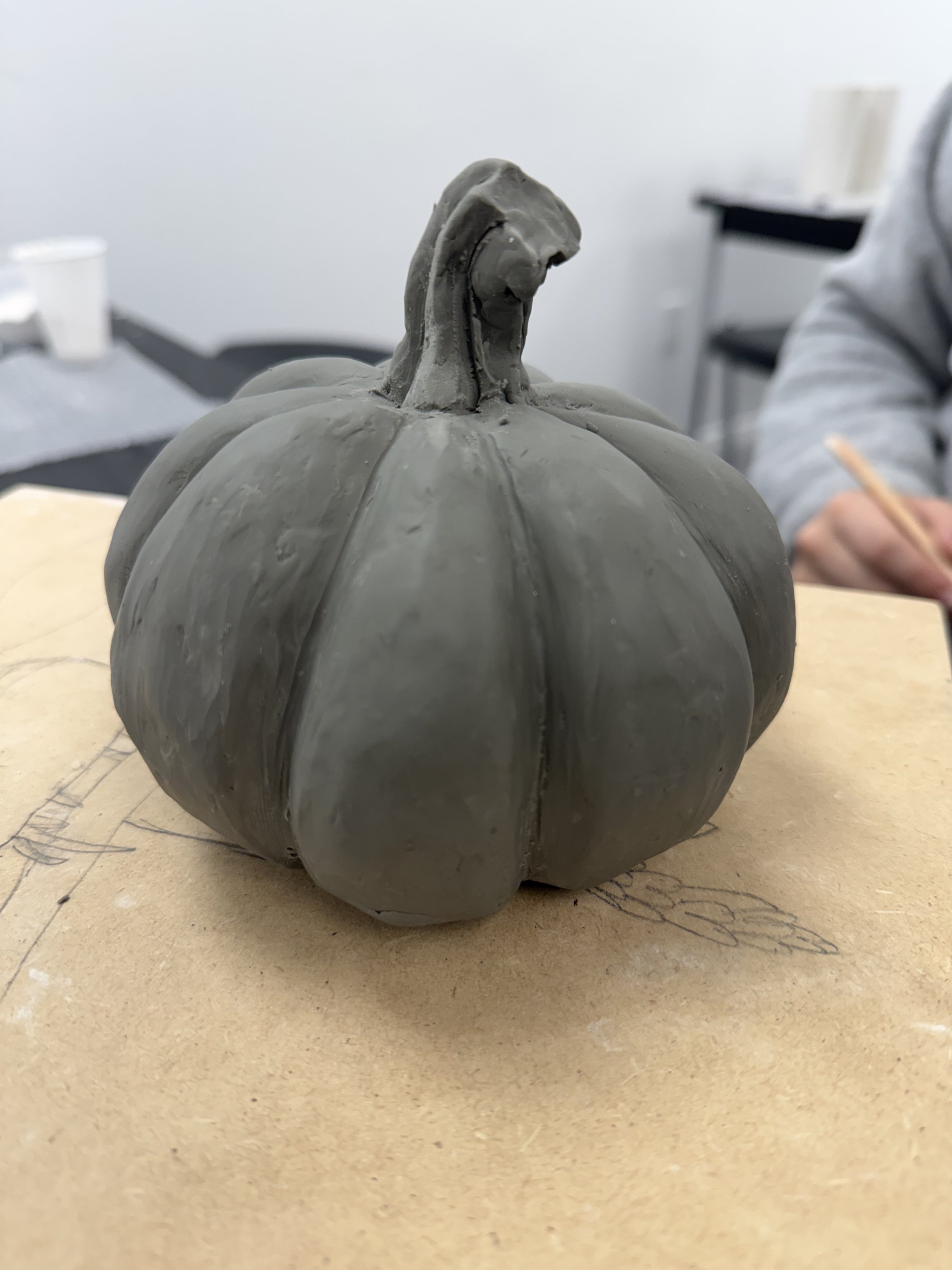 A gray clay sculpture of a pumpkin with prominent ridges and a stem on top, placed on a wooden surface.