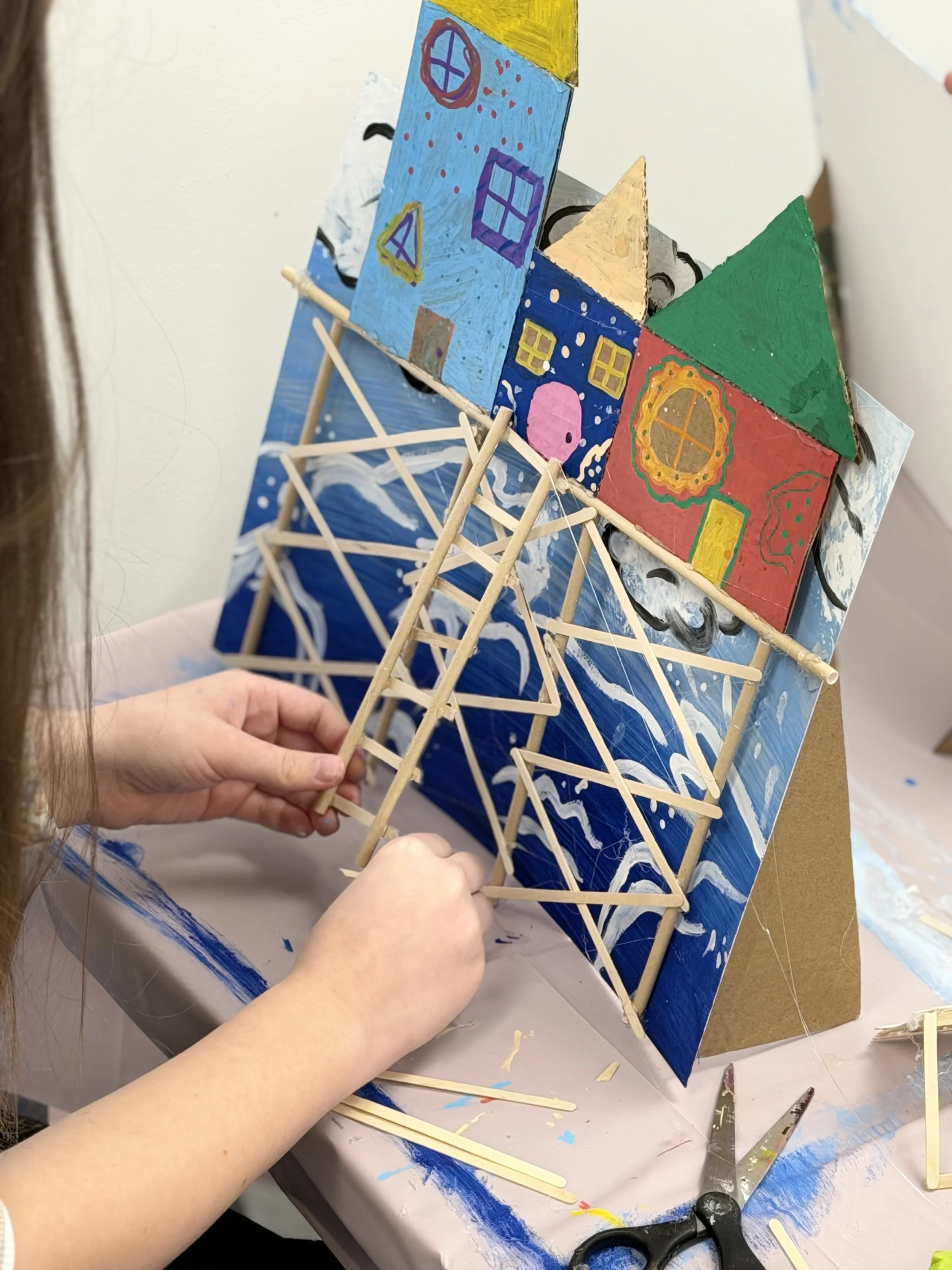 Child building a small wooden house and bridge with painted cardboard house backdrop.