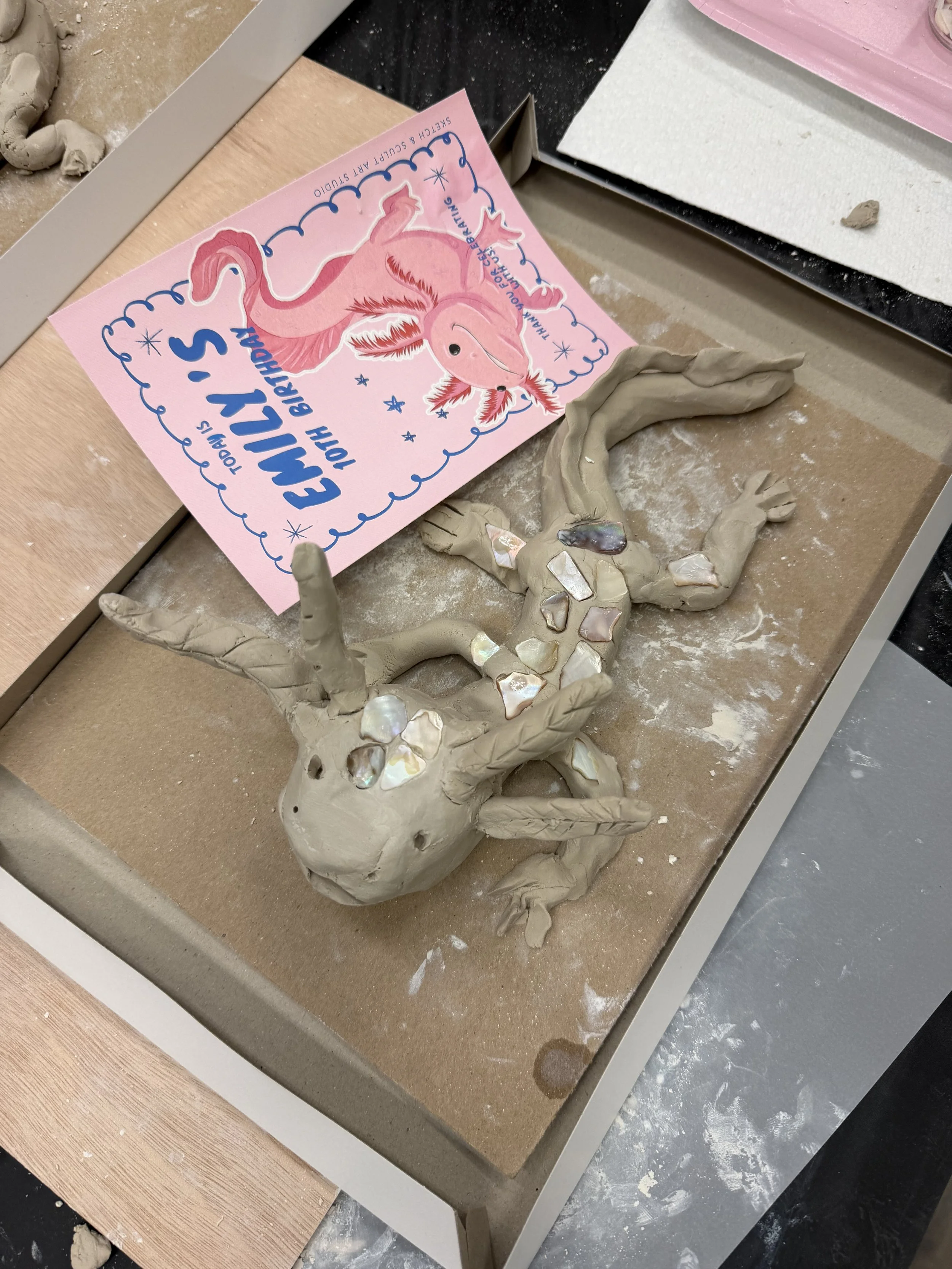 A clay sculpture of a lizard covered in shiny gemstones, placed on a workspace with a birthday card for Evelyn's 11th birthday featuring a pink unicorn, along with various art supplies.