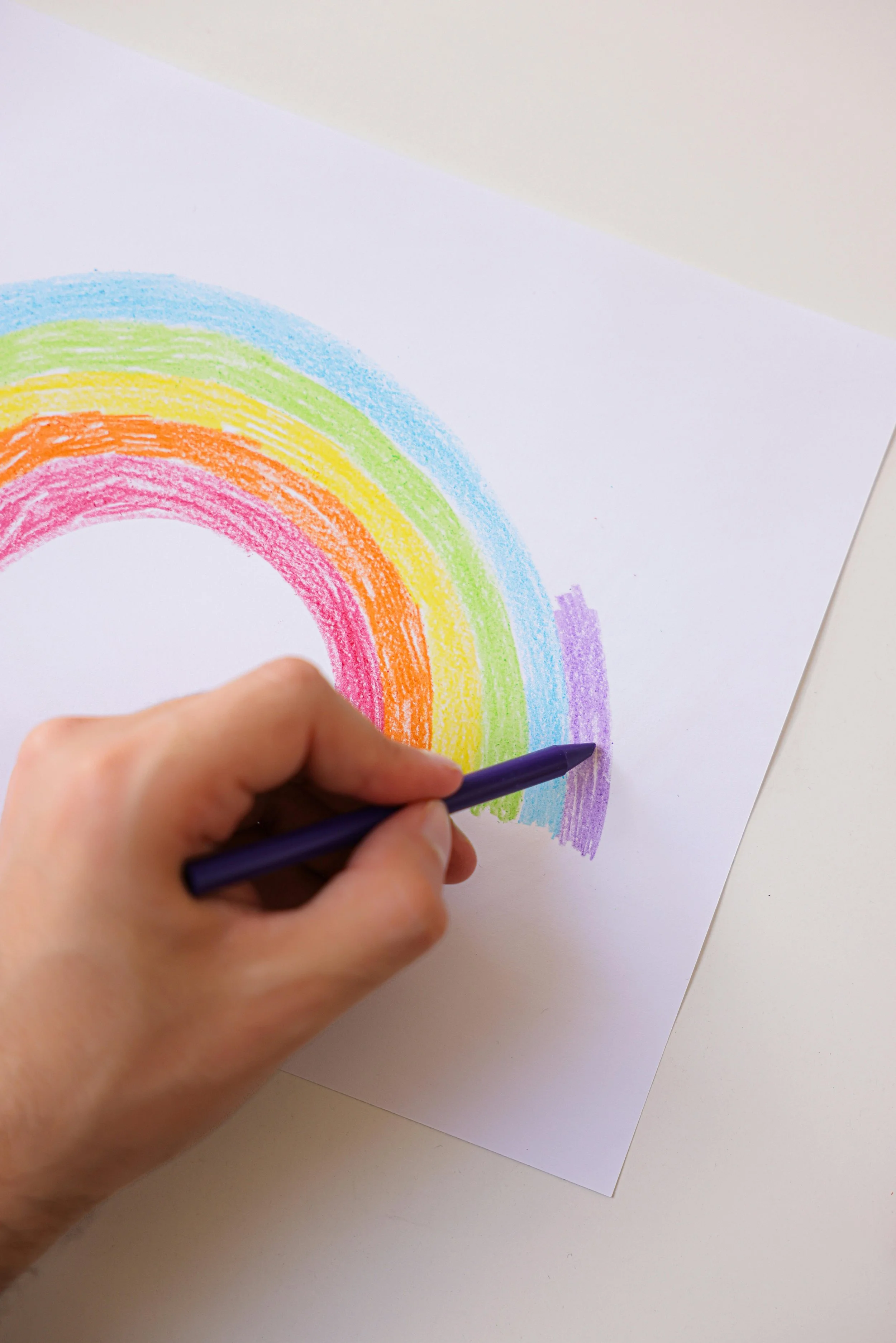 A person coloring a rainbow on a white sheet of paper with six different colored crayons.