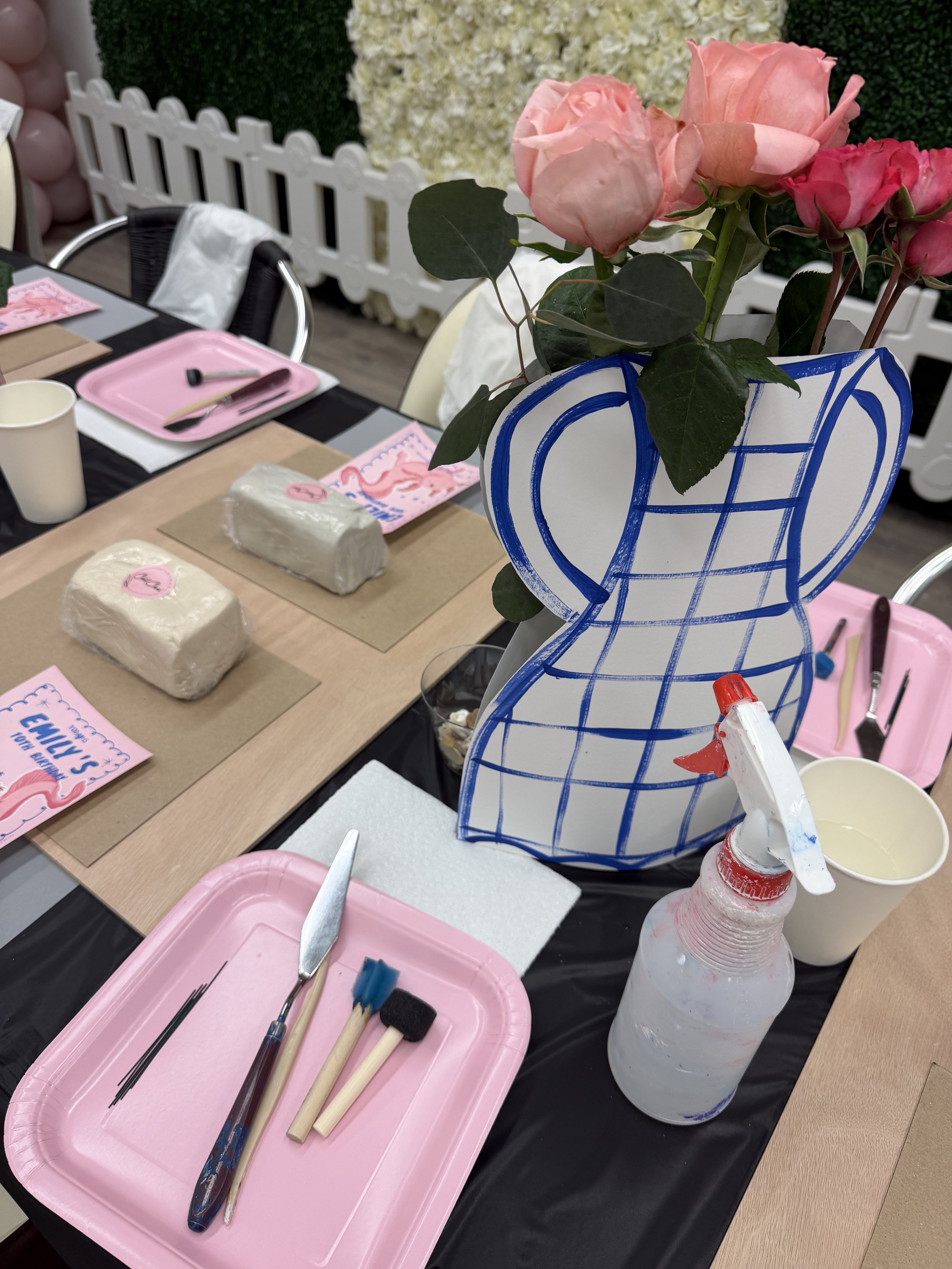 Decorative table with pink roses in a vase-shaped container, art supplies including brushes and tools, pink trays, and bags of modeling clay, set up for a painting or craft activity.