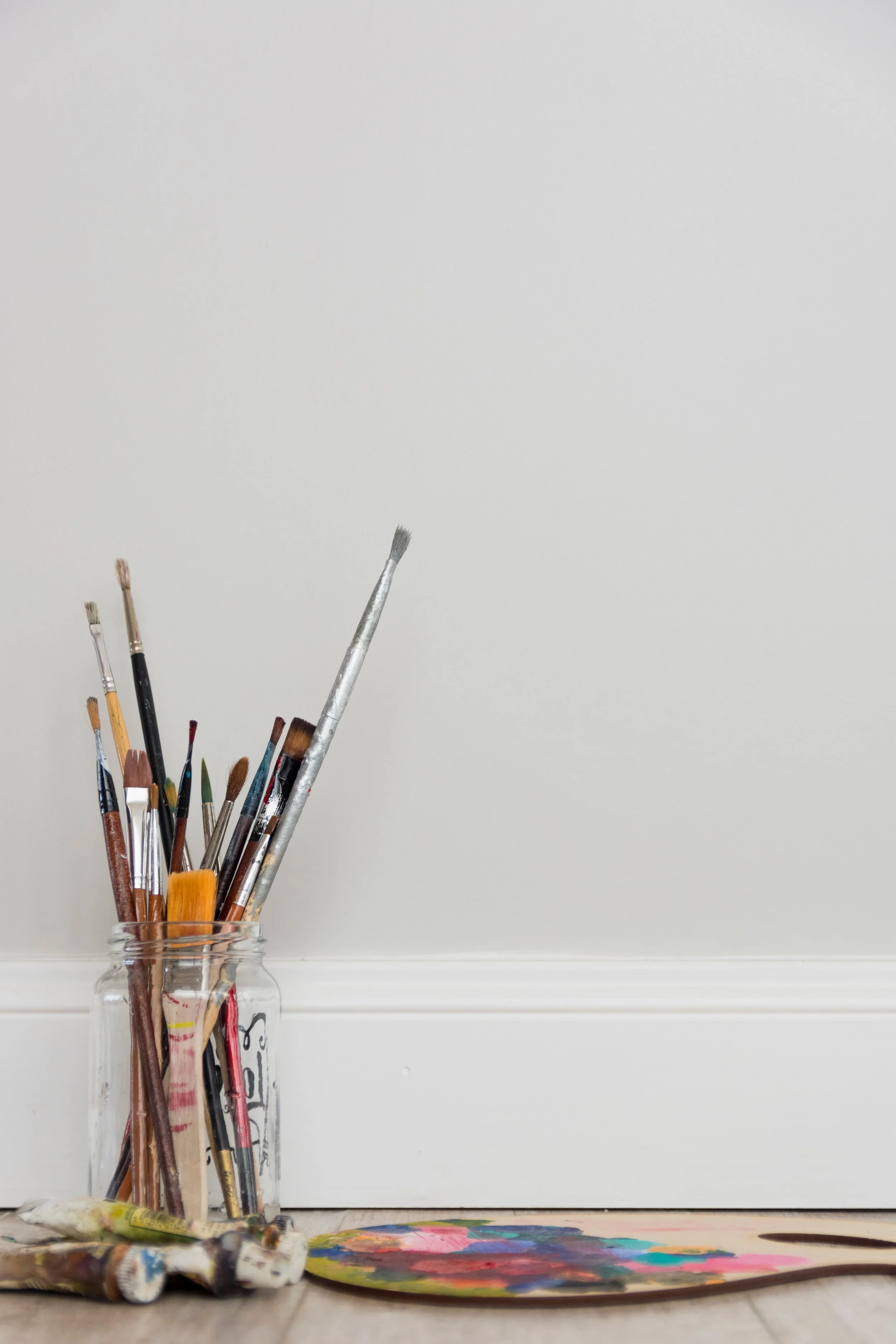 Paintbrushes in a glass jar, with a paint palette and tubes of paint on the floor, against a plain white wall.