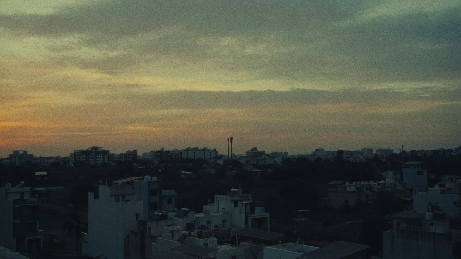 Cityscape at sunset with silhouettes of buildings and a cloudy sky.