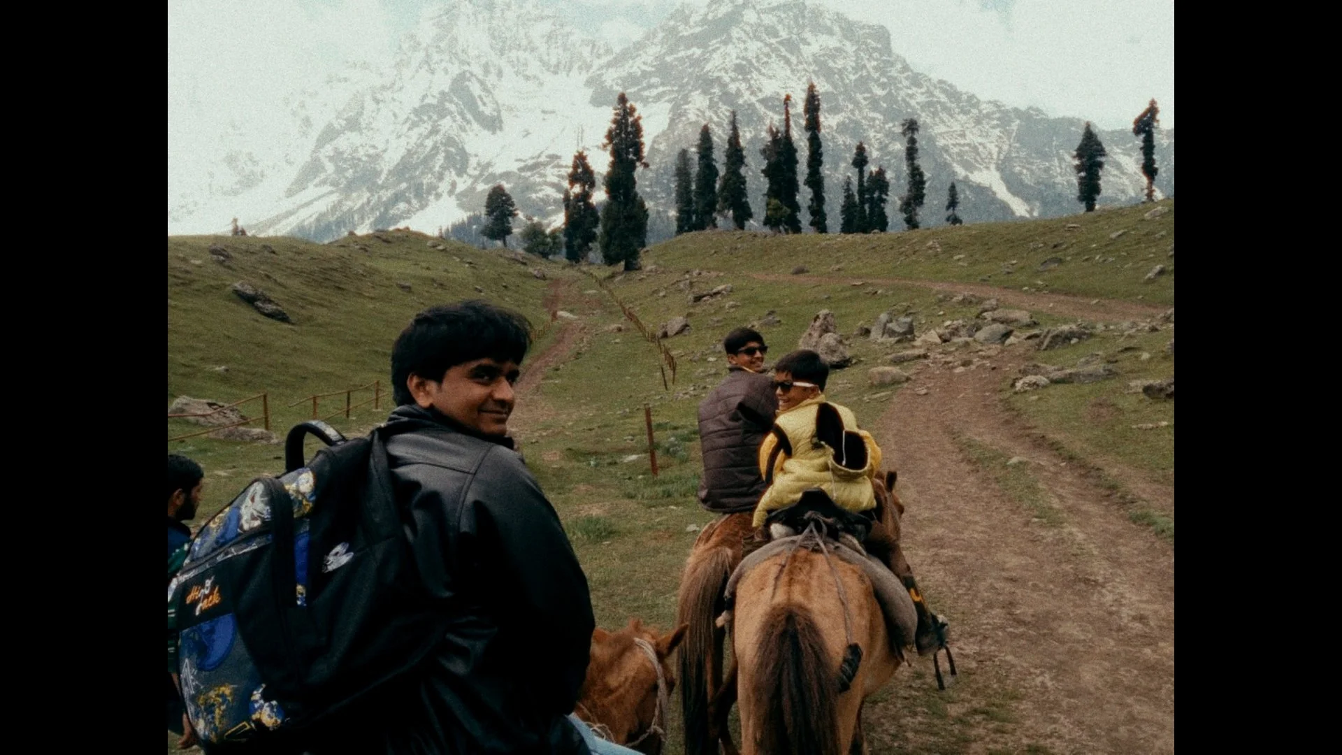 Two men riding camels on a dirt path in a mountainous area with snow-capped peaks and tall, thin trees in the background. One man is looking back, smiling, while the other is facing forward.