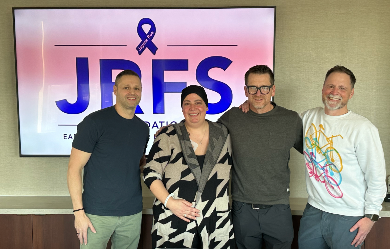 Four people smiling and standing in front of a large screen that reads 'JRFS Foundation' and has a purple awareness ribbon with the name Jayme Rae. The group is at a charity event.
