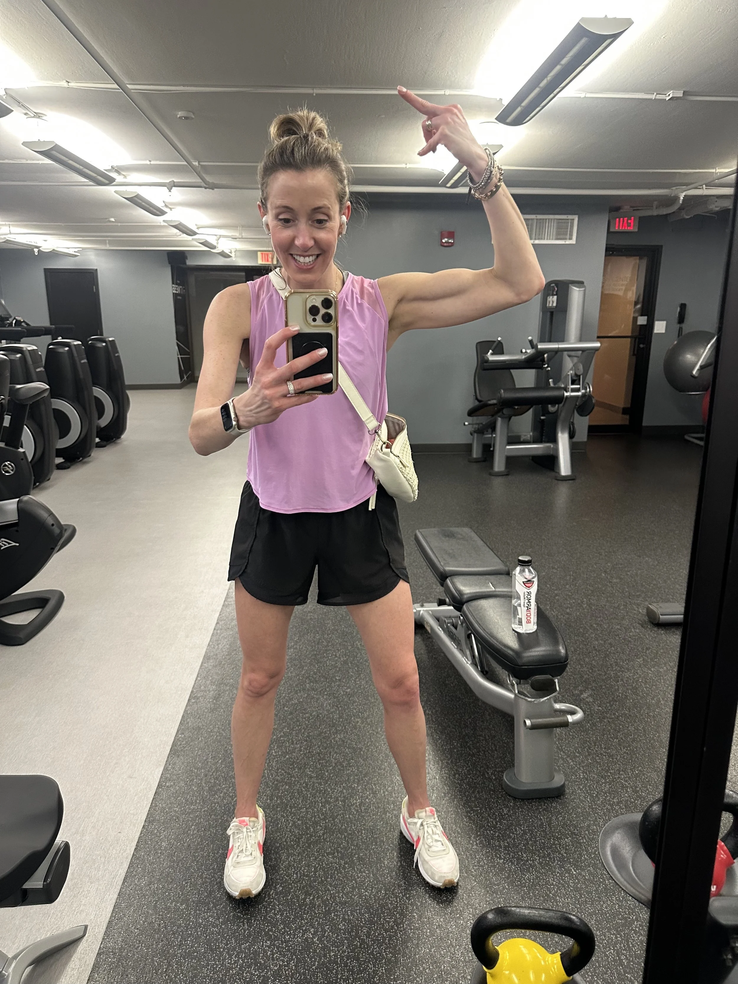 Jayme Forker Schwartz taking a mirror selfie in a gym. She is wearing a pink top, black shorts, and white sneakers. She is smiling, flexing her right arm, and has her hair tied up in a bun.