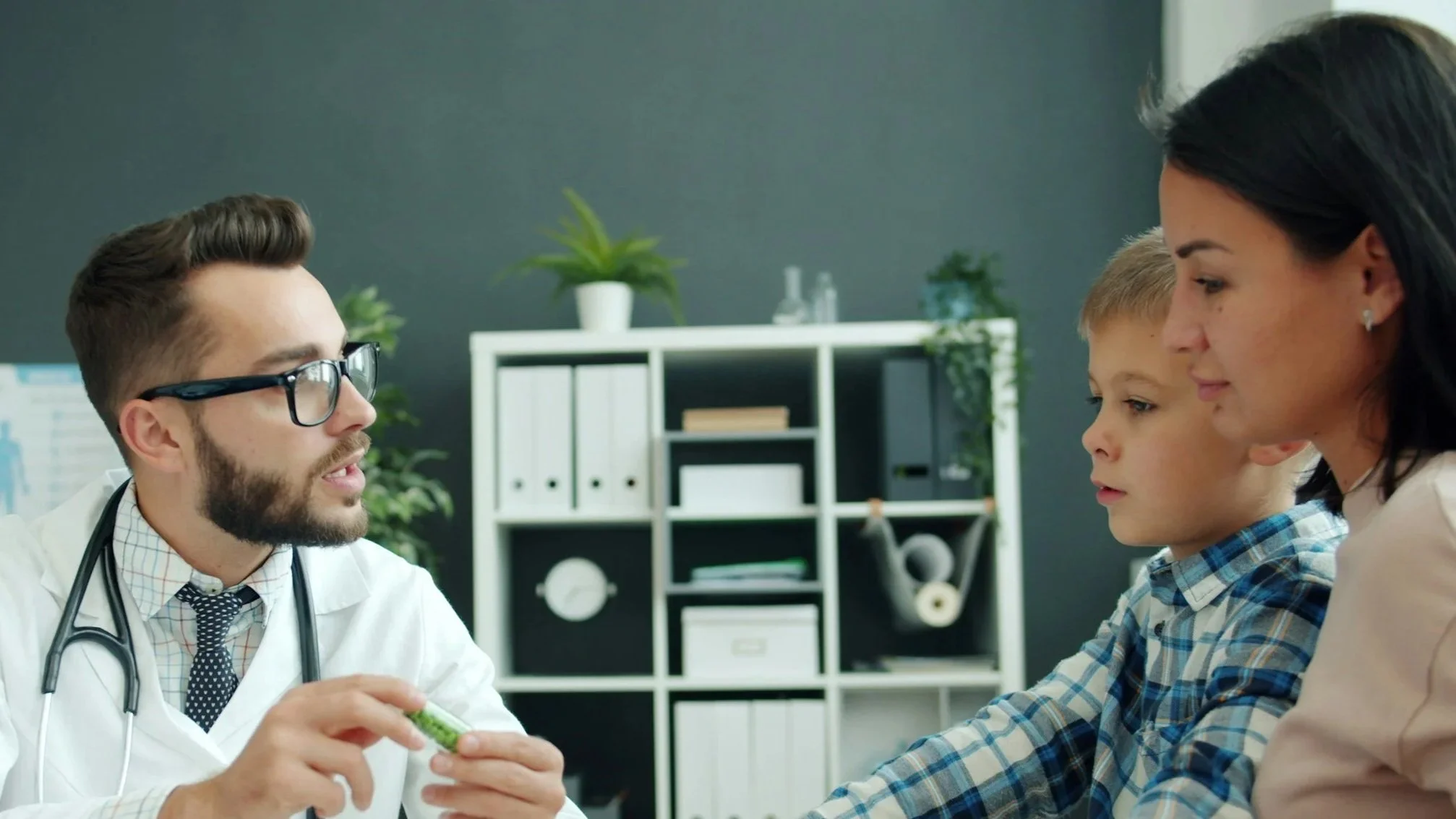 Doctor talking to a young boy and a woman in a medical office with a bookshelf and potted plants in the background.