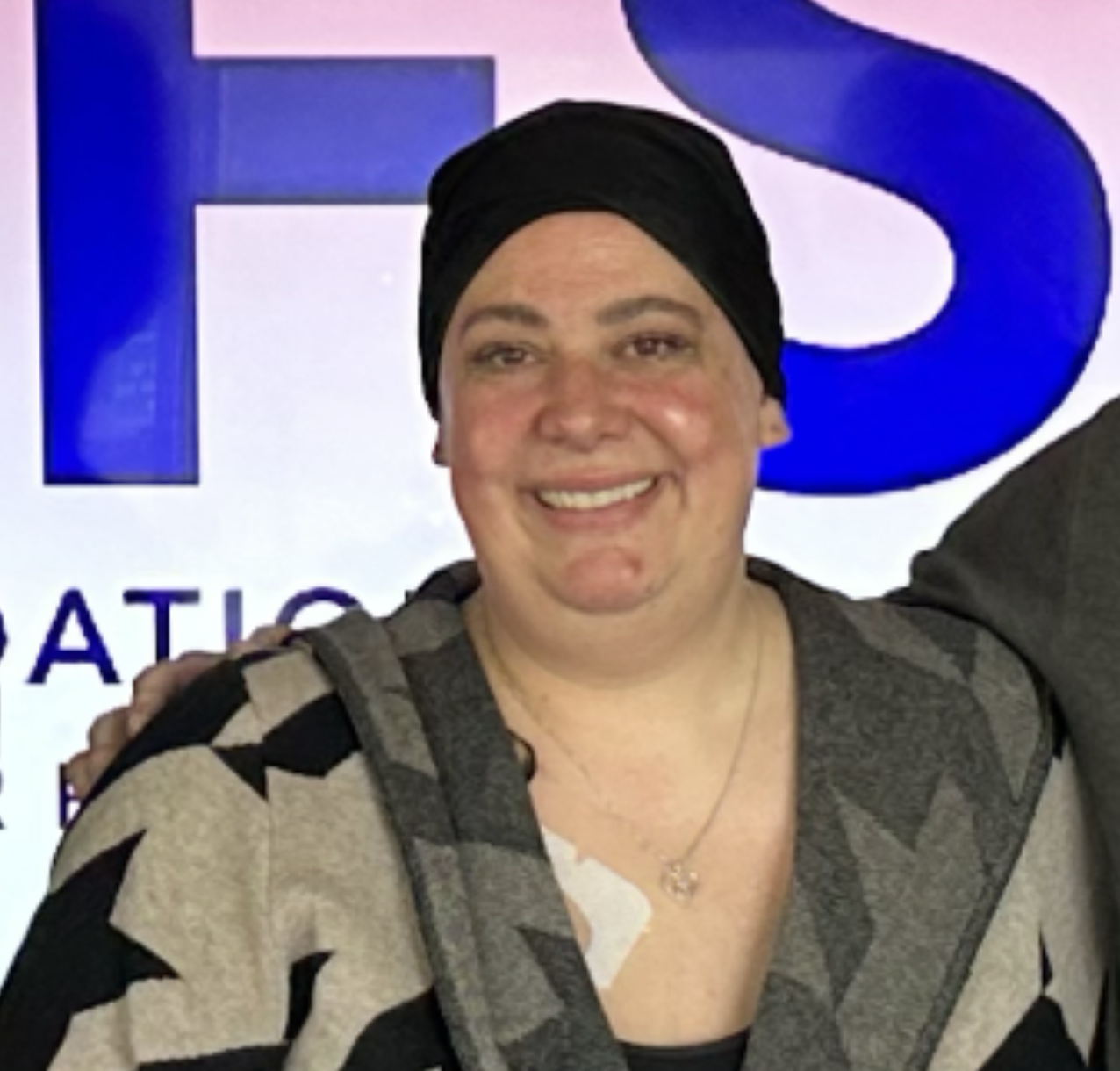 A smiling woman wearing a black headscarf and a hospital gown, standing in front of a partially visible purple and white JRFS Foundation sign.