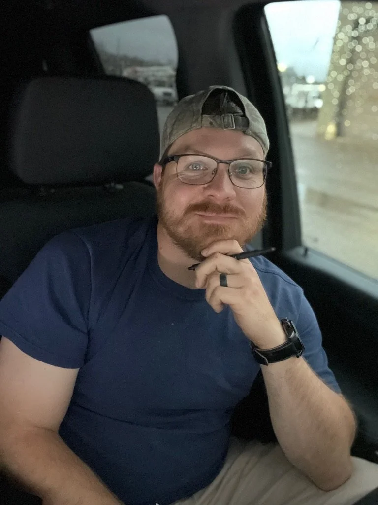 A man with glasses and a beard sitting inside a vehicle with a dark interior, wearing a grey backwards cap and a navy blue t-shirt, holding a pen or stylus near his chin, looking at the camera.