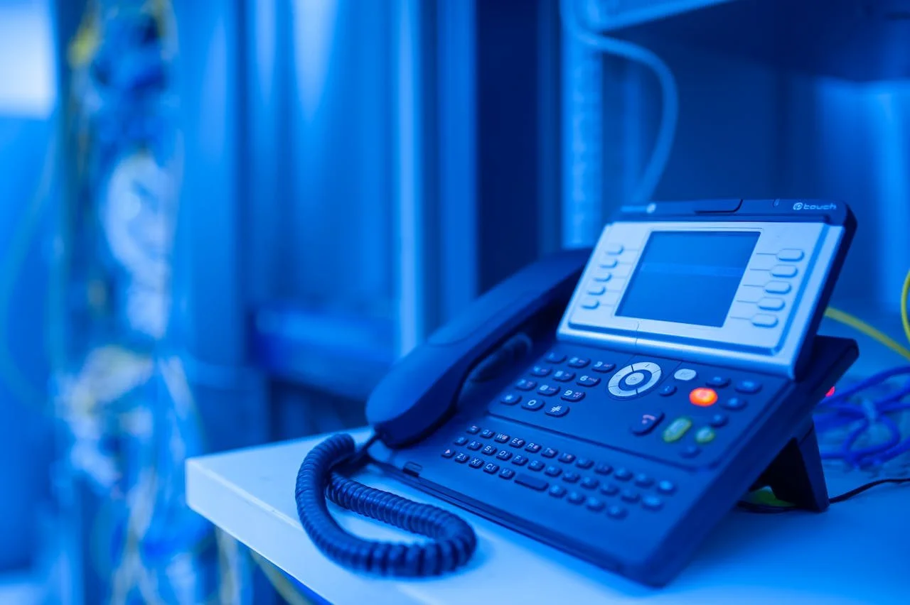 Office desk with a multi-line office phone, connected to a power source, on a white table, with a blurred background.
