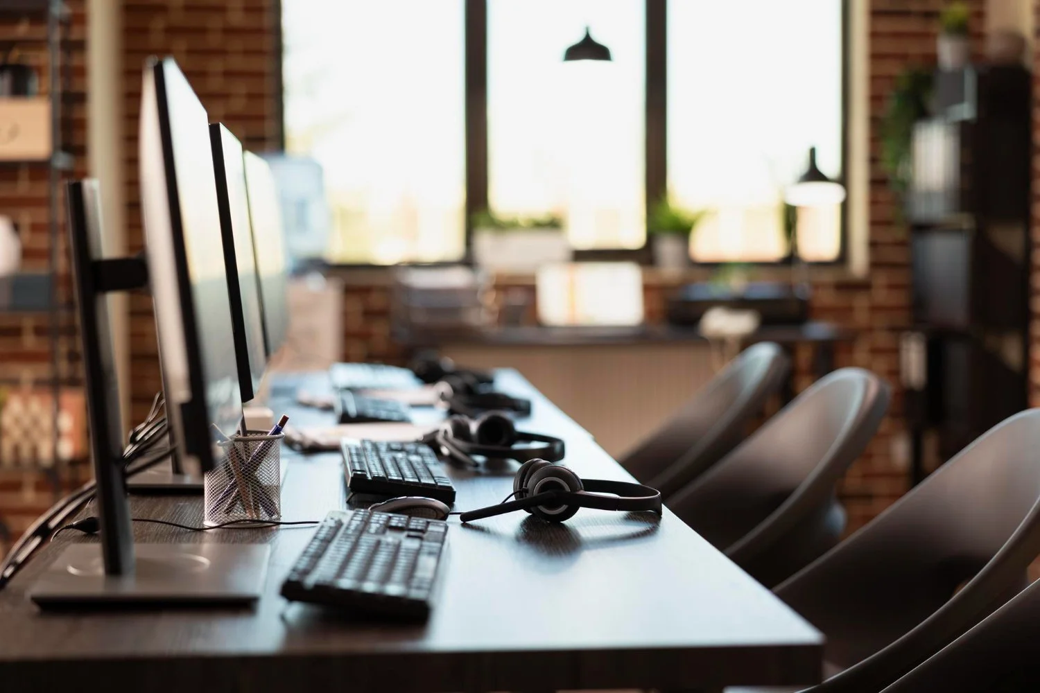 A modern office workspace with multiple computer monitors, keyboards, and headsets on a long wooden desk, with chairs arranged around it and a large window in the background.