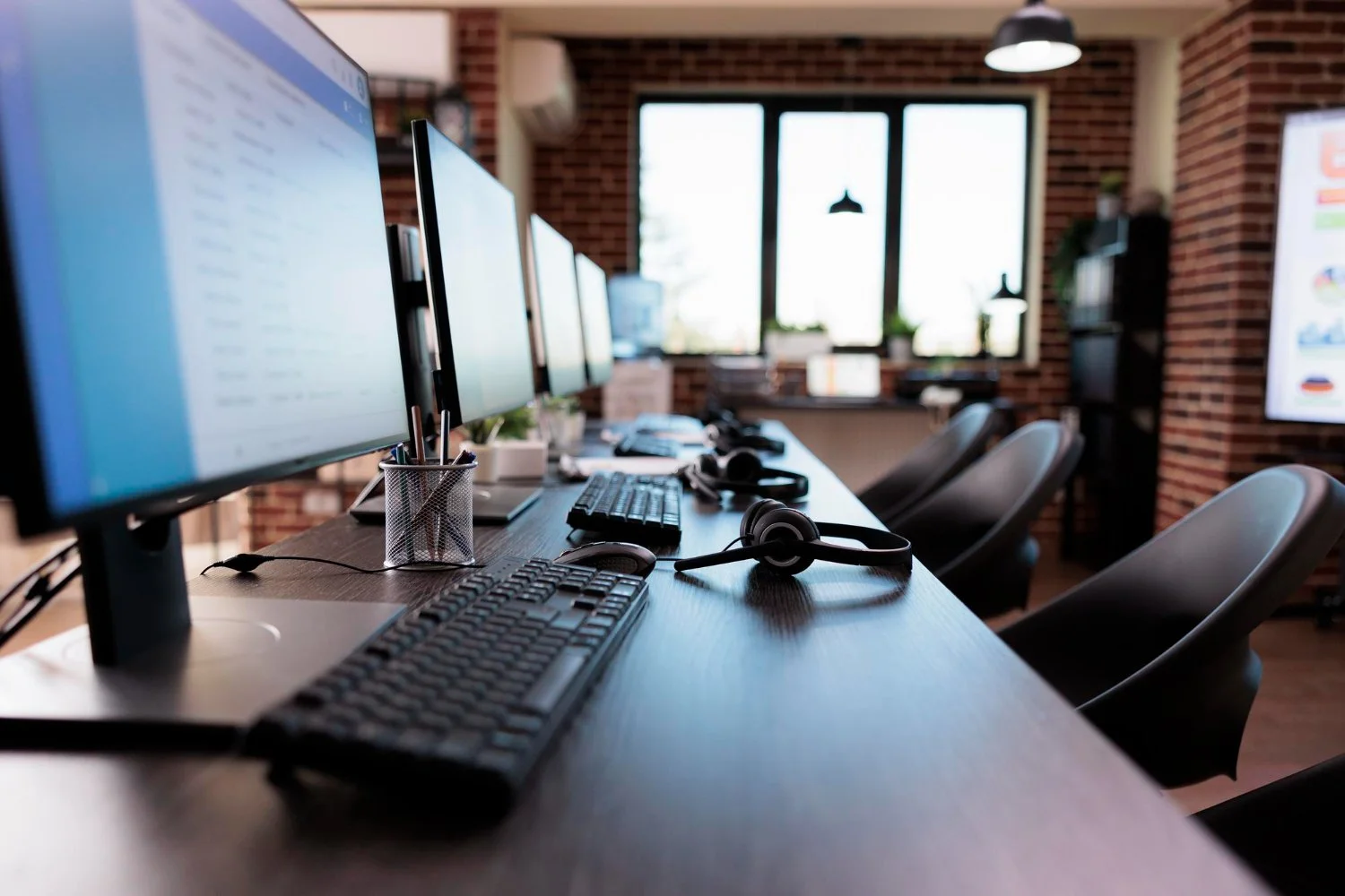 An office workspace with multiple computer monitors, keyboards, and headsets on a wooden desk, with chairs and a window in the background.