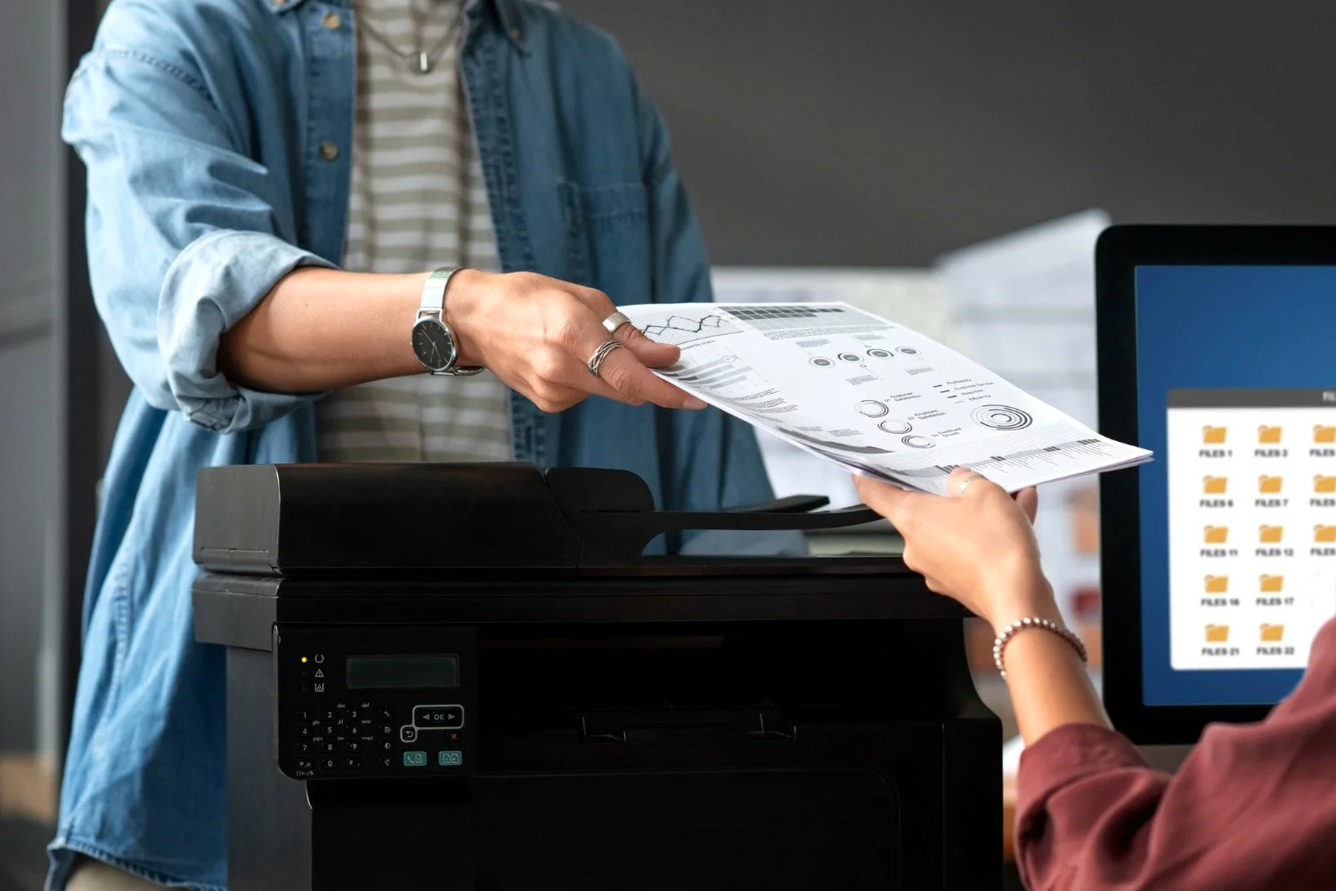 Person handing documents to another person at a photocopier in an office setting.