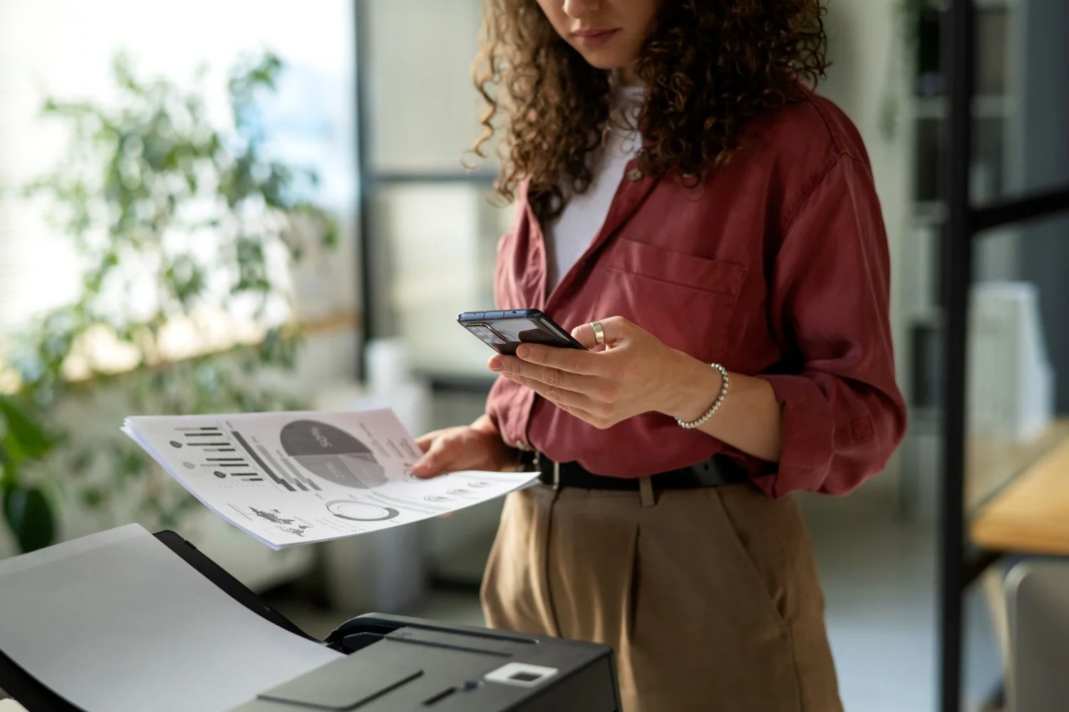 A woman with curly hair, wearing a red jacket and beige pants, is holding a smartphone in one hand and printed documents in the other, standing next to a photocopier in a modern office surrounded by green plants and glass walls.