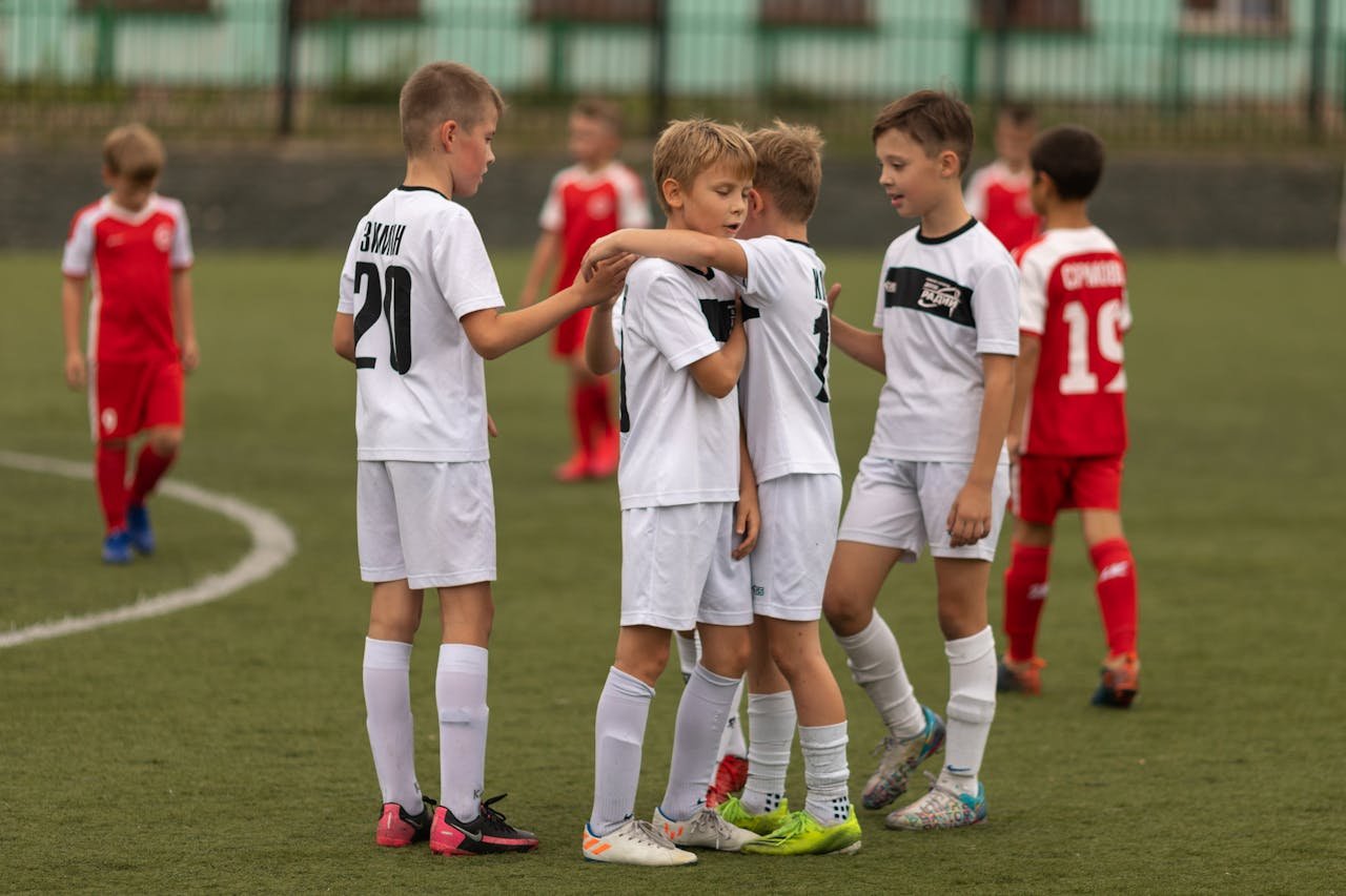 Young boys on a soccer field, some wearing white jerseys and others red, celebrating and comforting each other during a game.