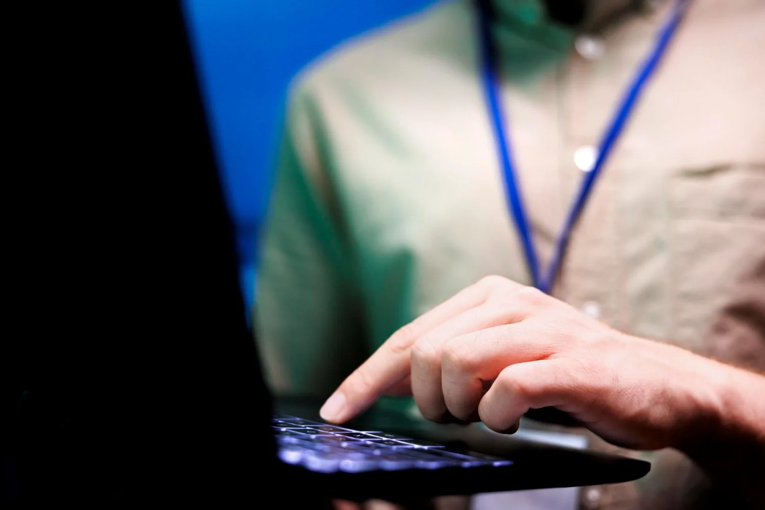 Close-up of a person's hand typing on a laptop keyboard, wearing a badge or lanyard around their neck.