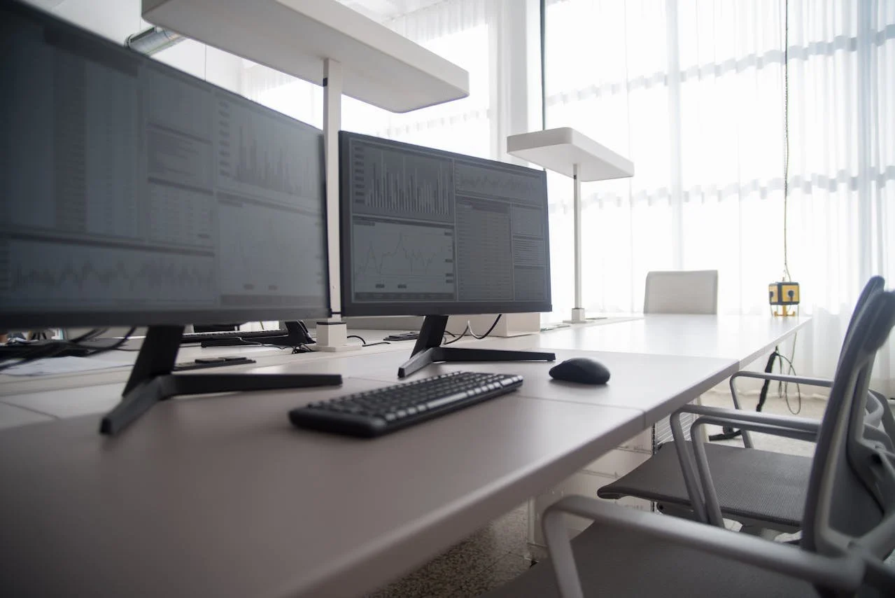 Empty modern office workspace with multiple computer monitors, keyboard, mouse, chairs, and large windows with curtains letting in natural light.