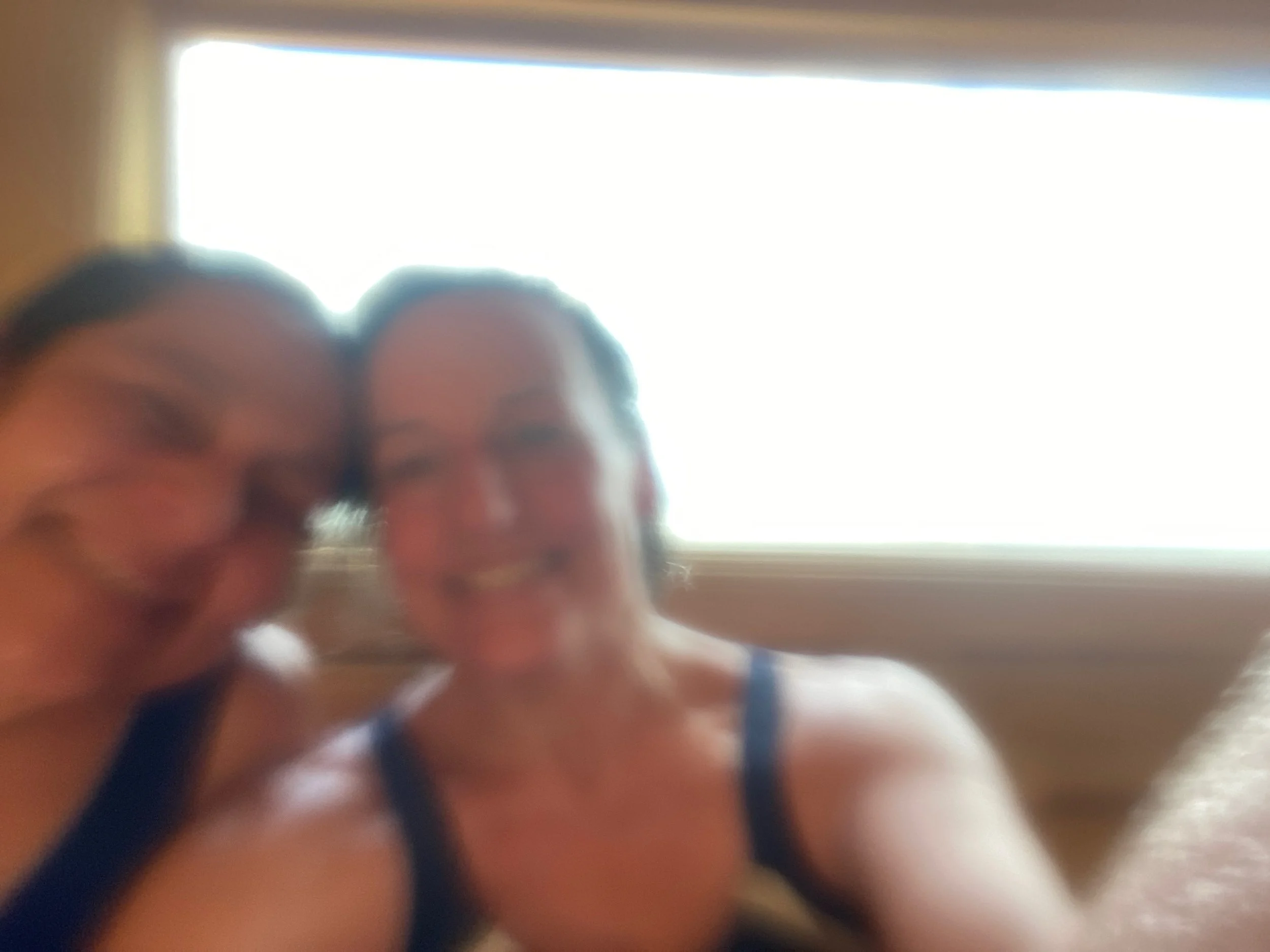 Two women smiling and taking a selfie in front of a bright window in a steam-filled sauna.