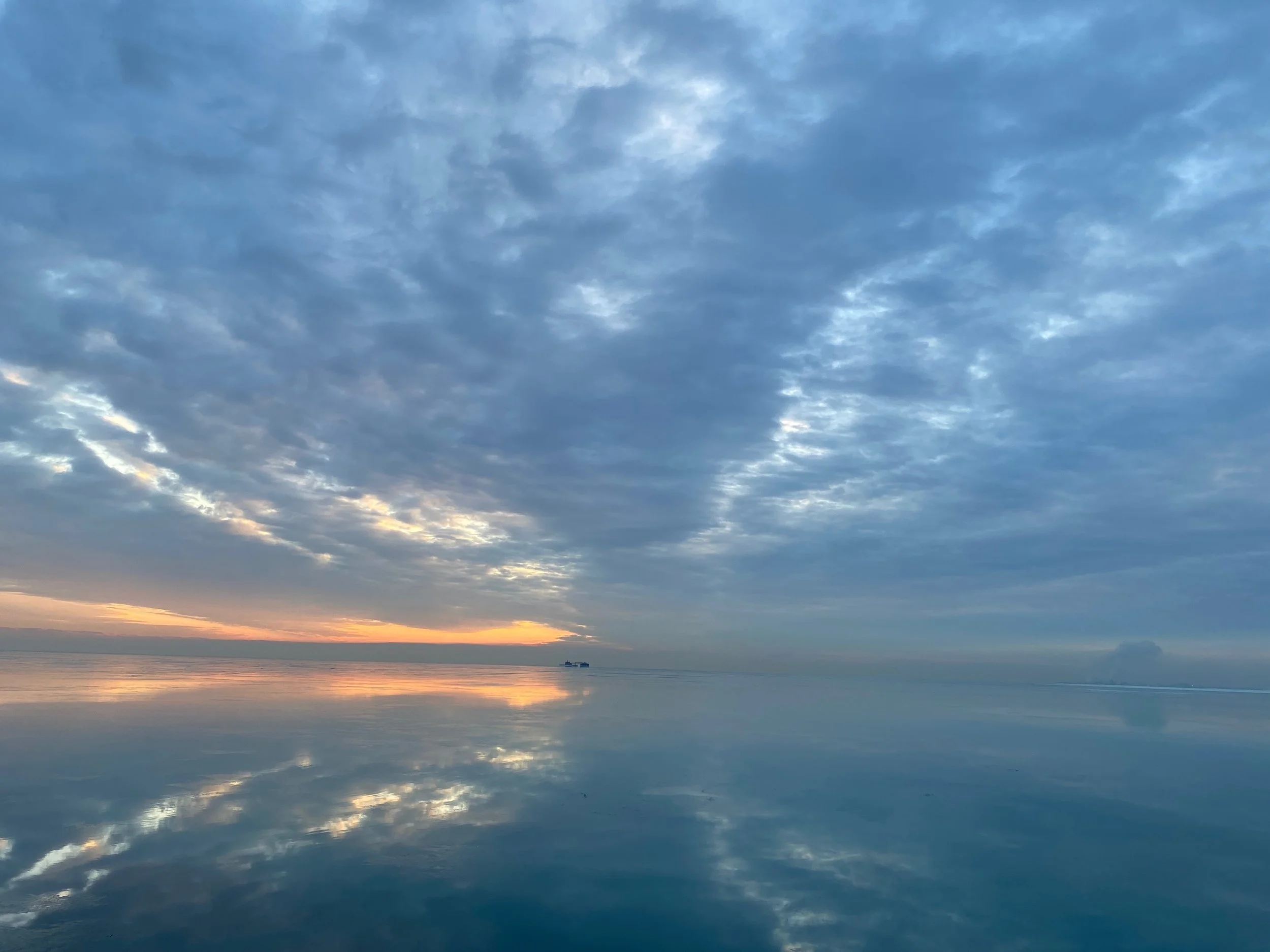 Calm view of Lake Michigan reflecting a cloudy sky at sunrise or sunset, with a small boat on the horizon.