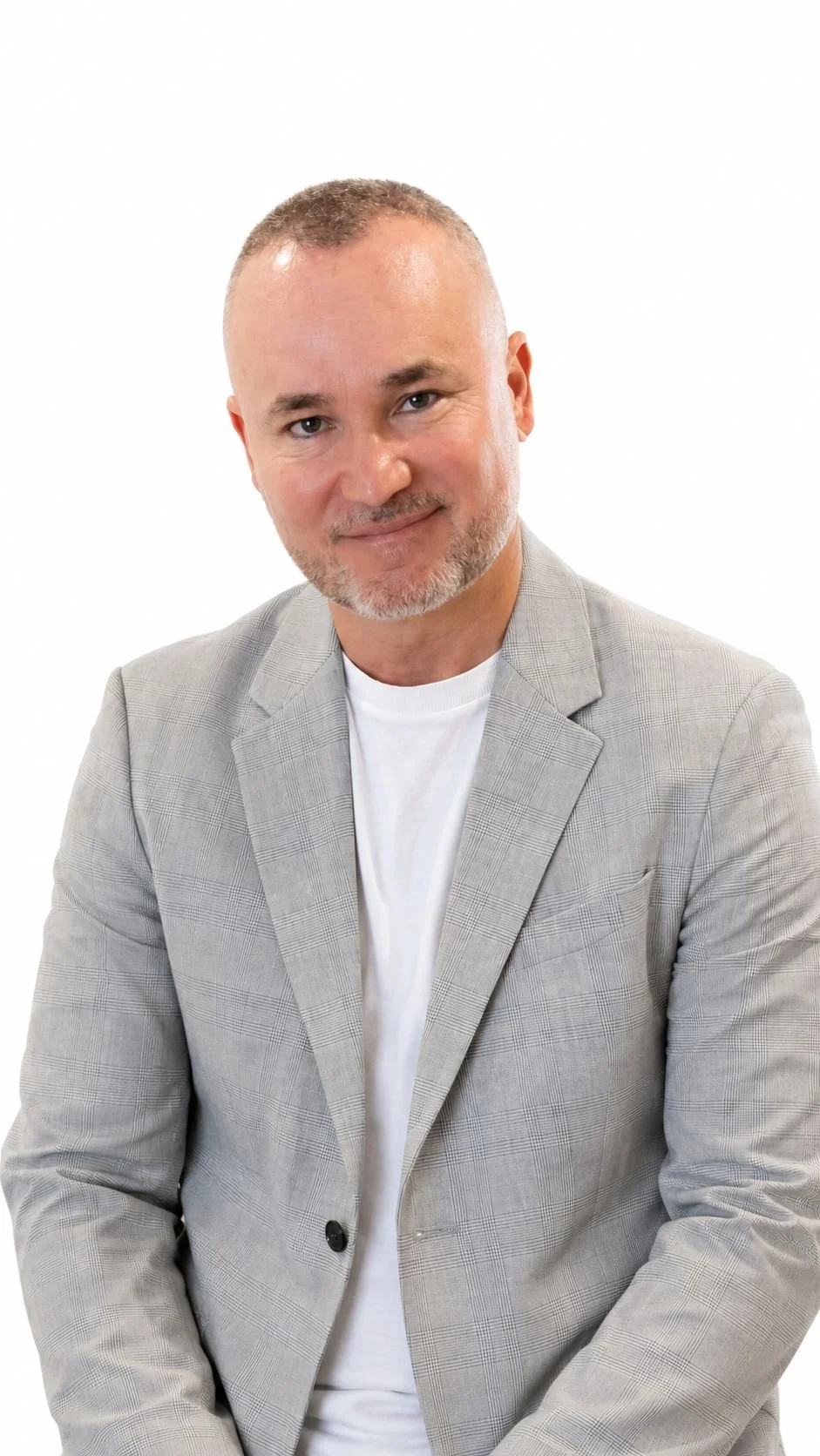 A middle-aged man with short gray hair, a trimmed beard, and a friendly smile, wearing a light gray blazer over a white t-shirt, against a plain white background.