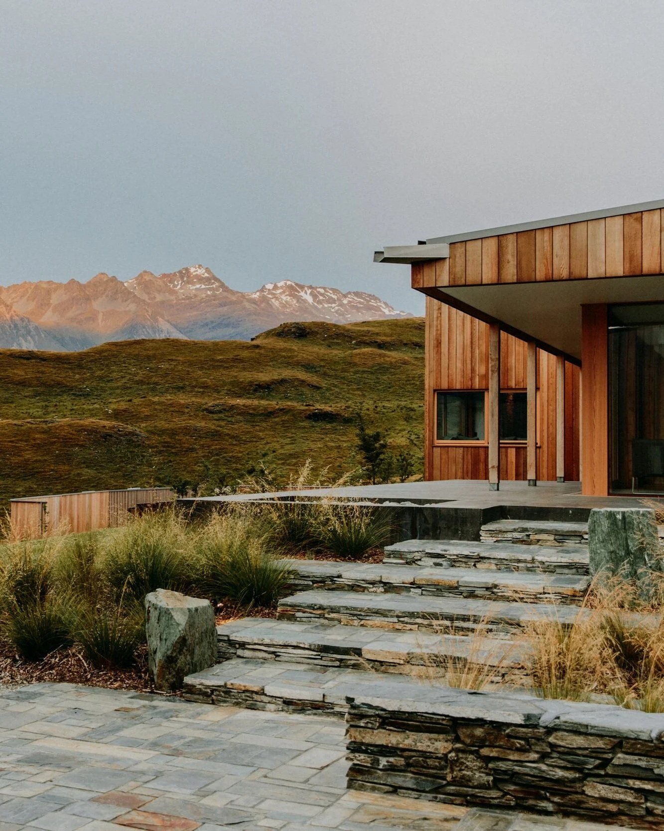 Modern house with wooden exterior, stone steps, and mountain landscape in the background.