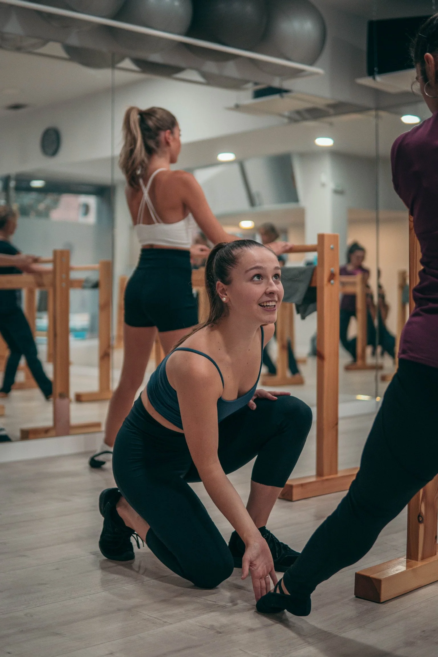 A young woman in workout clothes squats on the floor during a fitness class, smiling and interacting with an instructor in a gym with mirror walls.