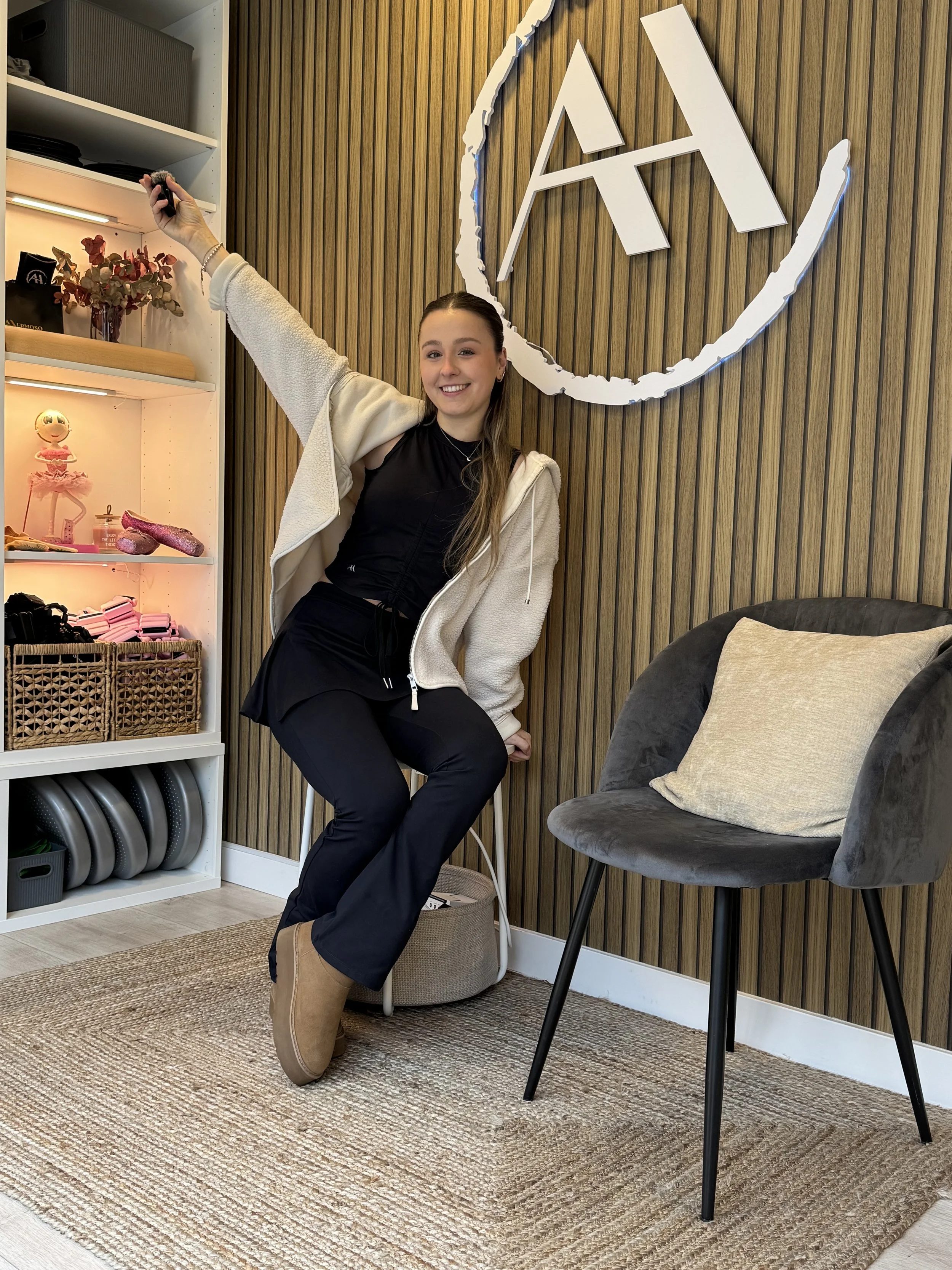 A young woman in black pants and tan boots smiling and waving with her arm raised, sitting on a stool in a modern room with wooden wall paneling and a large 'AI' logo.