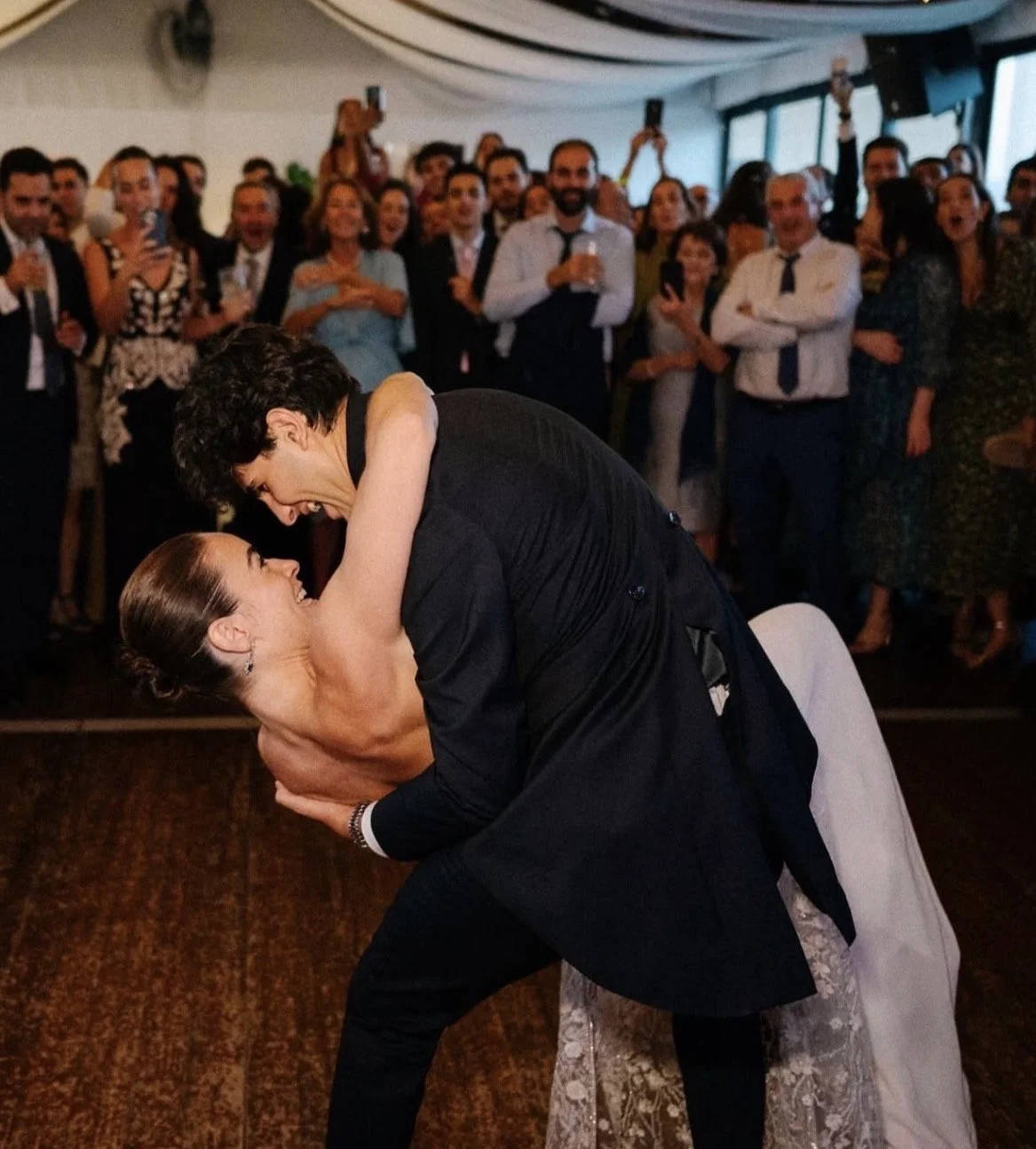 Pareja bailando en una boda, el hombre levanta a la mujer en una postura de danza, mientras la multitud observa y captura el momento con celulares en un espacio cerrado con luz natural.