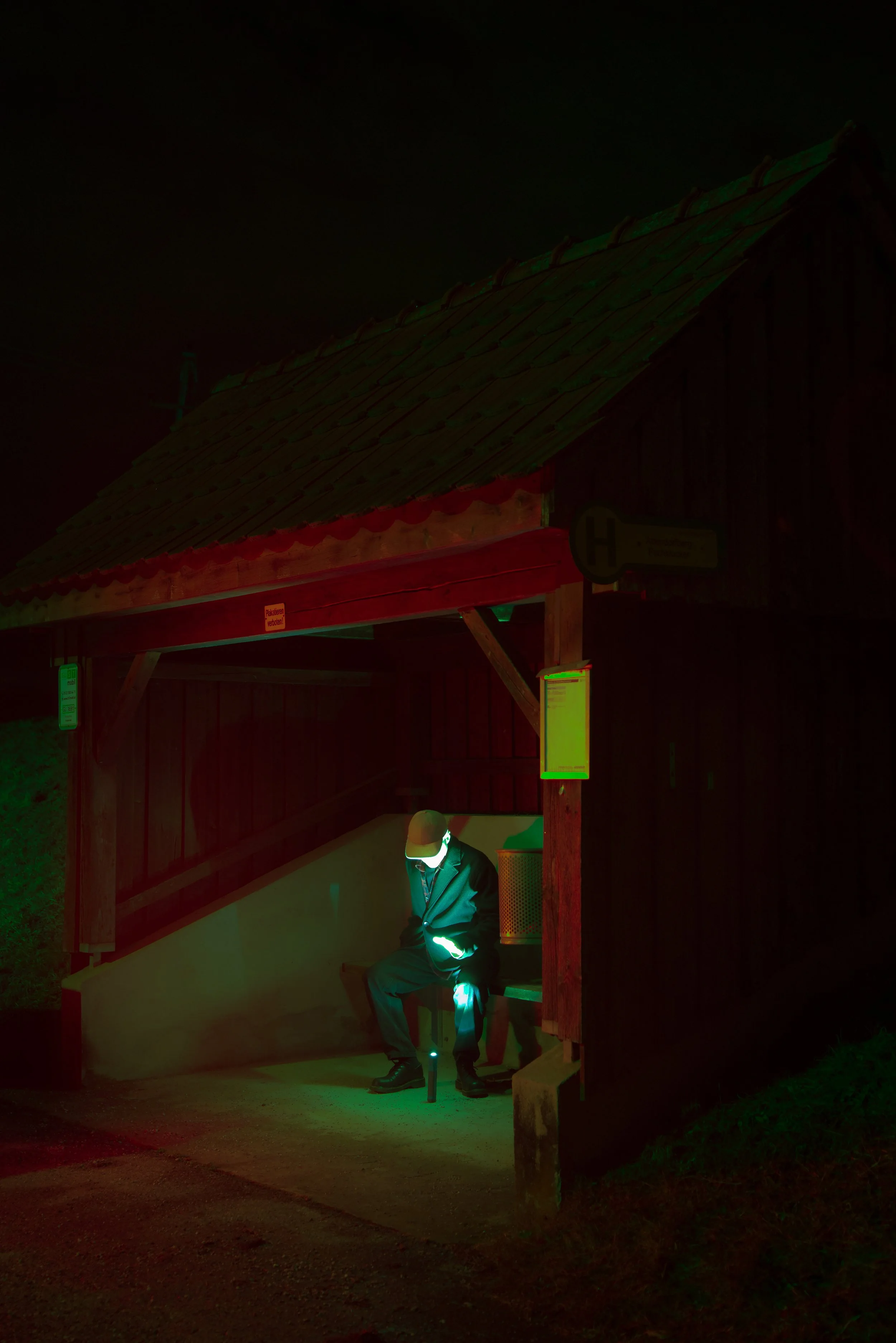 A person sitting on a bench at a bus stop shelter, illuminated by a greenish light, with darkness surrounding the shelter.
