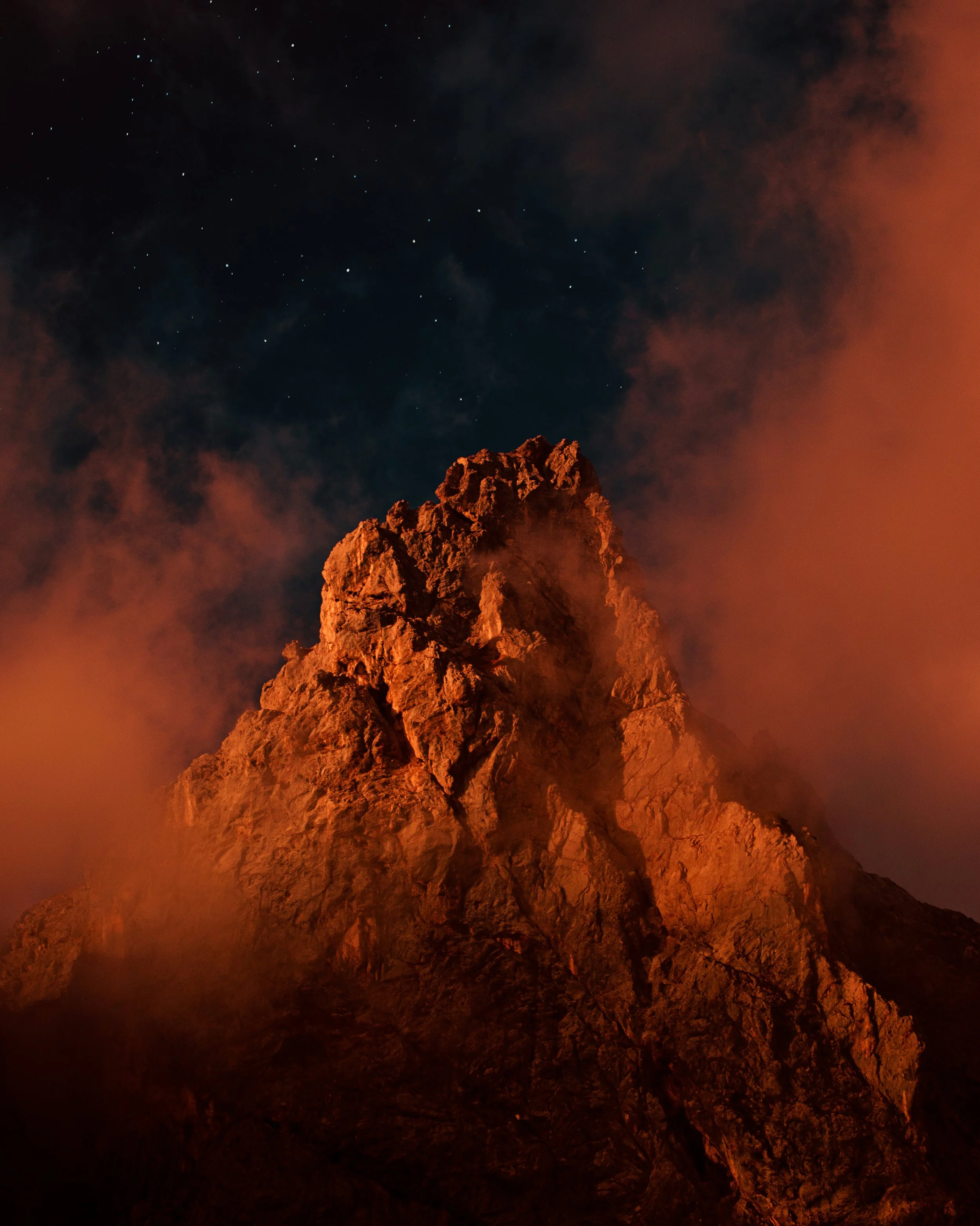Nighttime view of a rugged mountain peak illuminated with orange-red light, with clouds surrounding it and stars visible in the clear sky above.