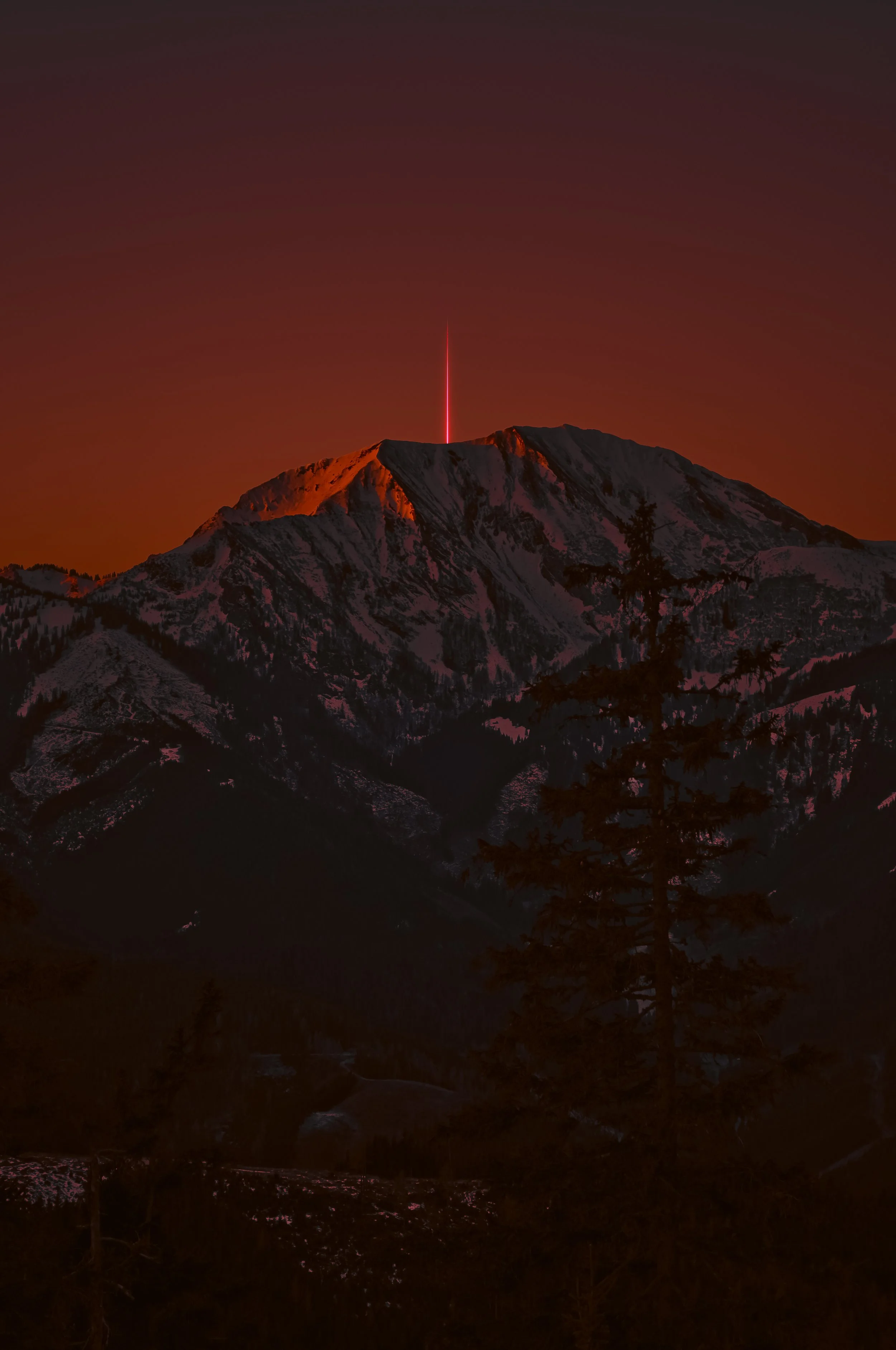 Snow-capped mountain with a bright red beam of light shooting into the sky at sunset or twilight.