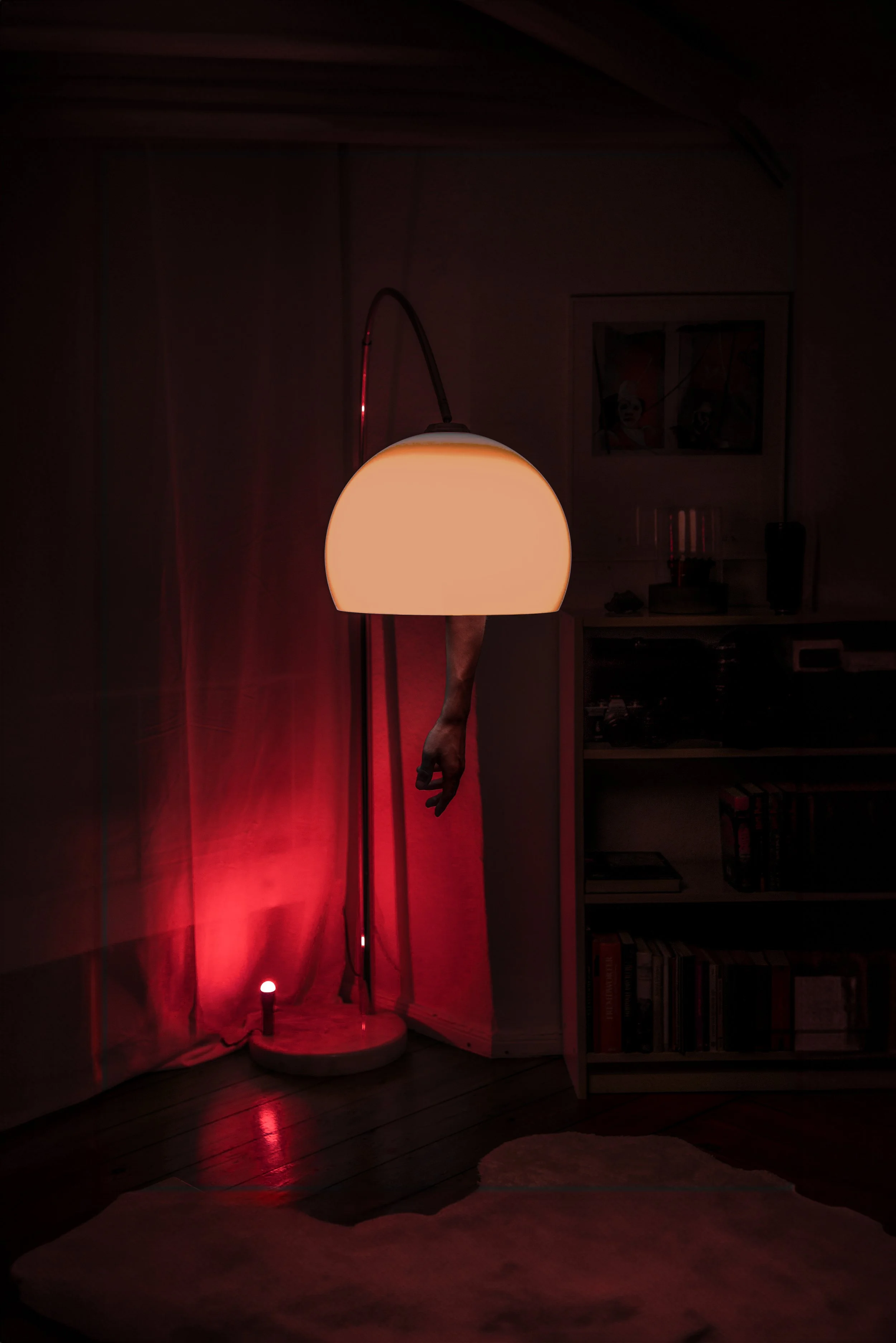 A dimly lit room with a floor lamp that has a white, dome-shaped shade. A human hand is hanging from behind the lamp, and red lighting illuminates the wall behind it. There is a bookshelf on the right side and a white rug on the floor.