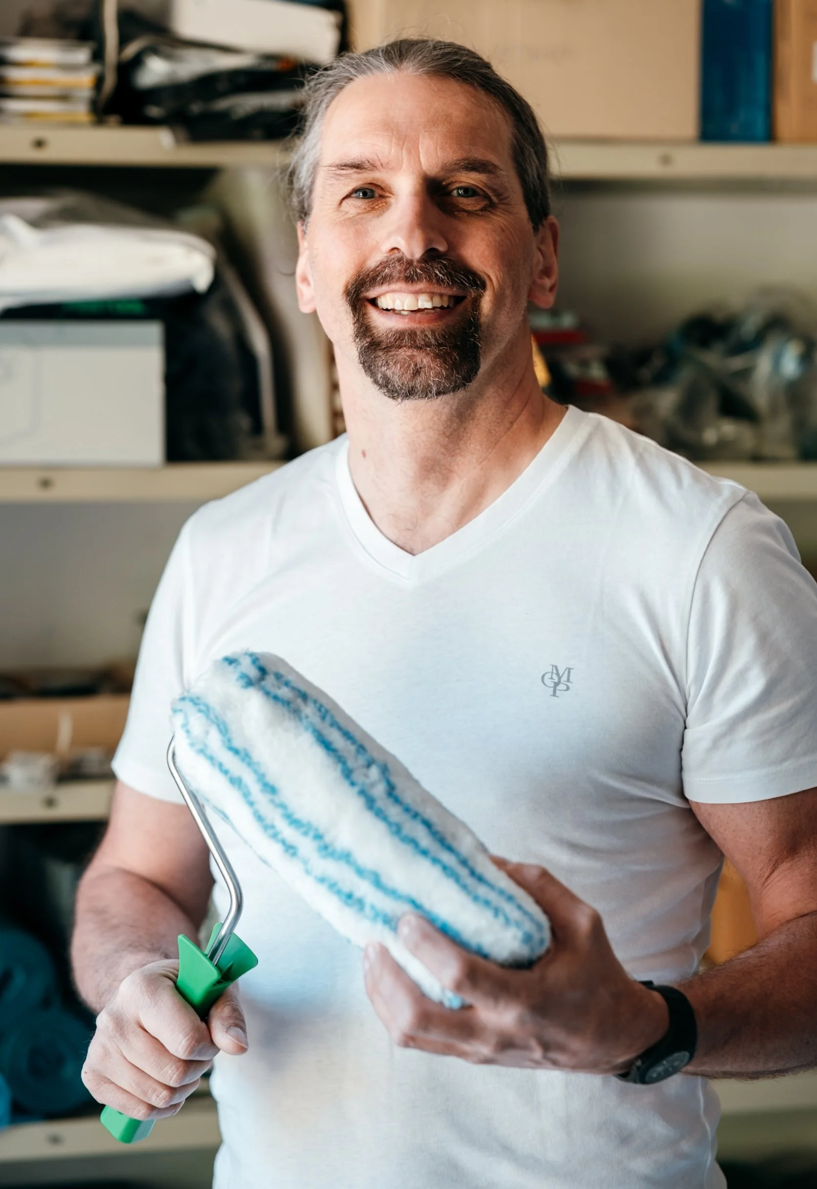 Ein Mann mit grauem Haar und Bart, der ein weißes T-Shirt trägt, hält eine Farbrolle und einen Pinsel, während er vor einem Regal mit Werkzeugen und Materialien steht.