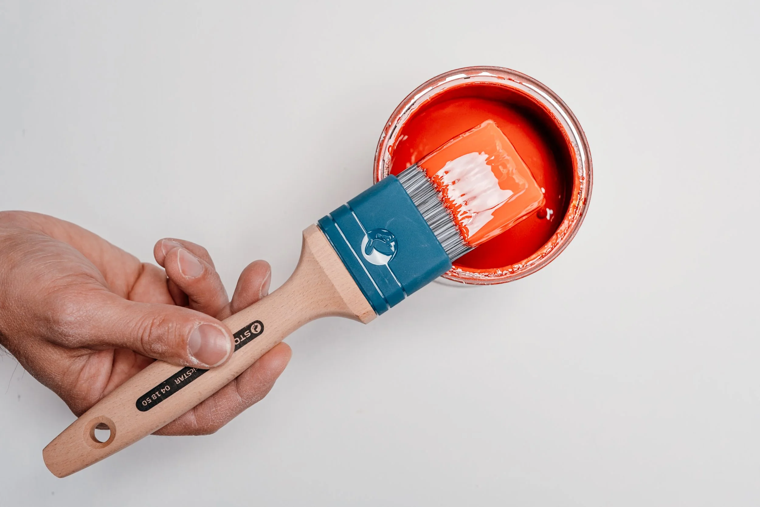 Hand hält einen Pinsel, der in eine Dose mit roter Farbe taucht, auf weißem Hintergrund.