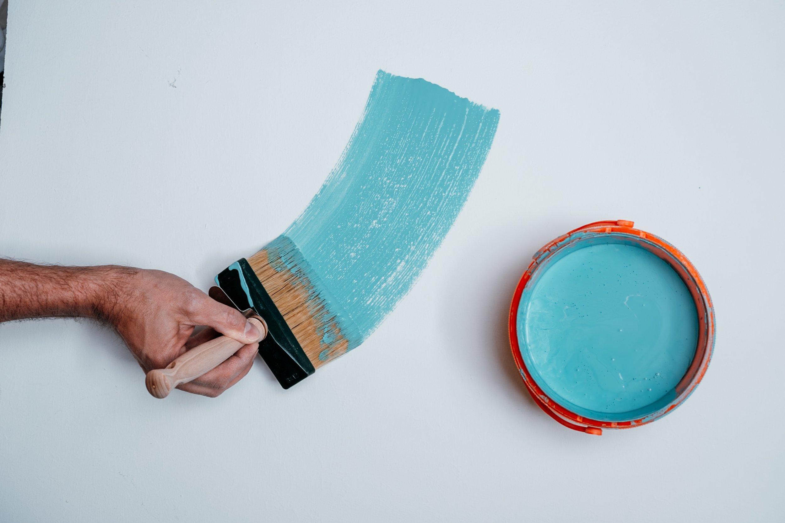 Person beim Streichen einer weißen Wand mit blauer Farbe, Pinsel in der Hand, Farbkübel auf einer weißen Oberfläche