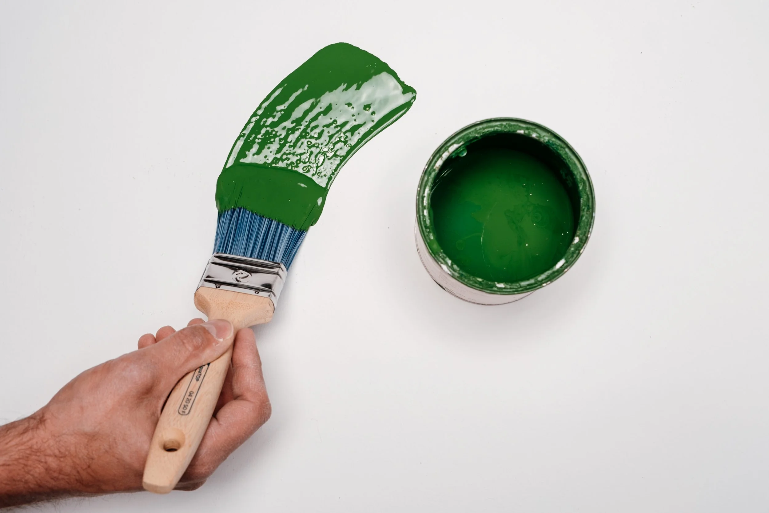 Hand hält einen Pinsel, der grüne Farbe aufträgt. Es gibt einen offenen Eimer mit grüner Farbe auf einer weißen Oberfläche.