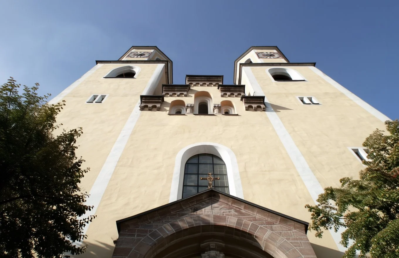 Aufnahme von unten auf eine kirchliche Fassade mit zwei hohen Türmen und einer kleinen Dachgiebel mit Kreuz, im Sonnenlicht, im Blauhimmel und umgeben von Bäumen.